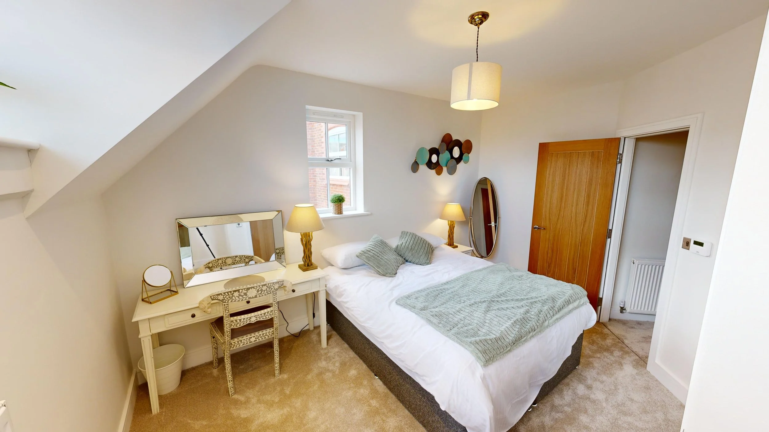 A cozy bedroom with a sloped ceiling, featuring a bed with light green bedding, bedside lamps, a mirror, a wooden wardrobe, and decorative wall art.