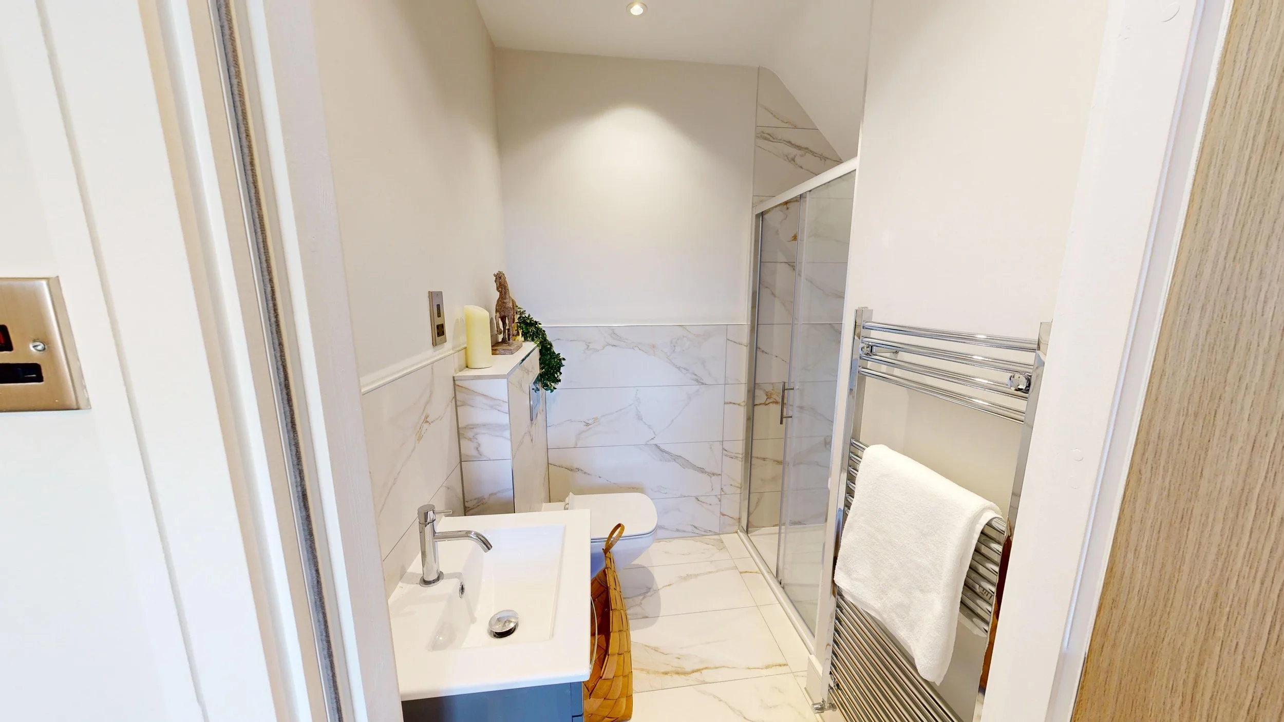 Modern small bathroom with marble tiles, a glass-enclosed shower, a wall-mounted white toilet, a small white sink with a chrome faucet, a wicker basket, and a towel rack with a white towel.