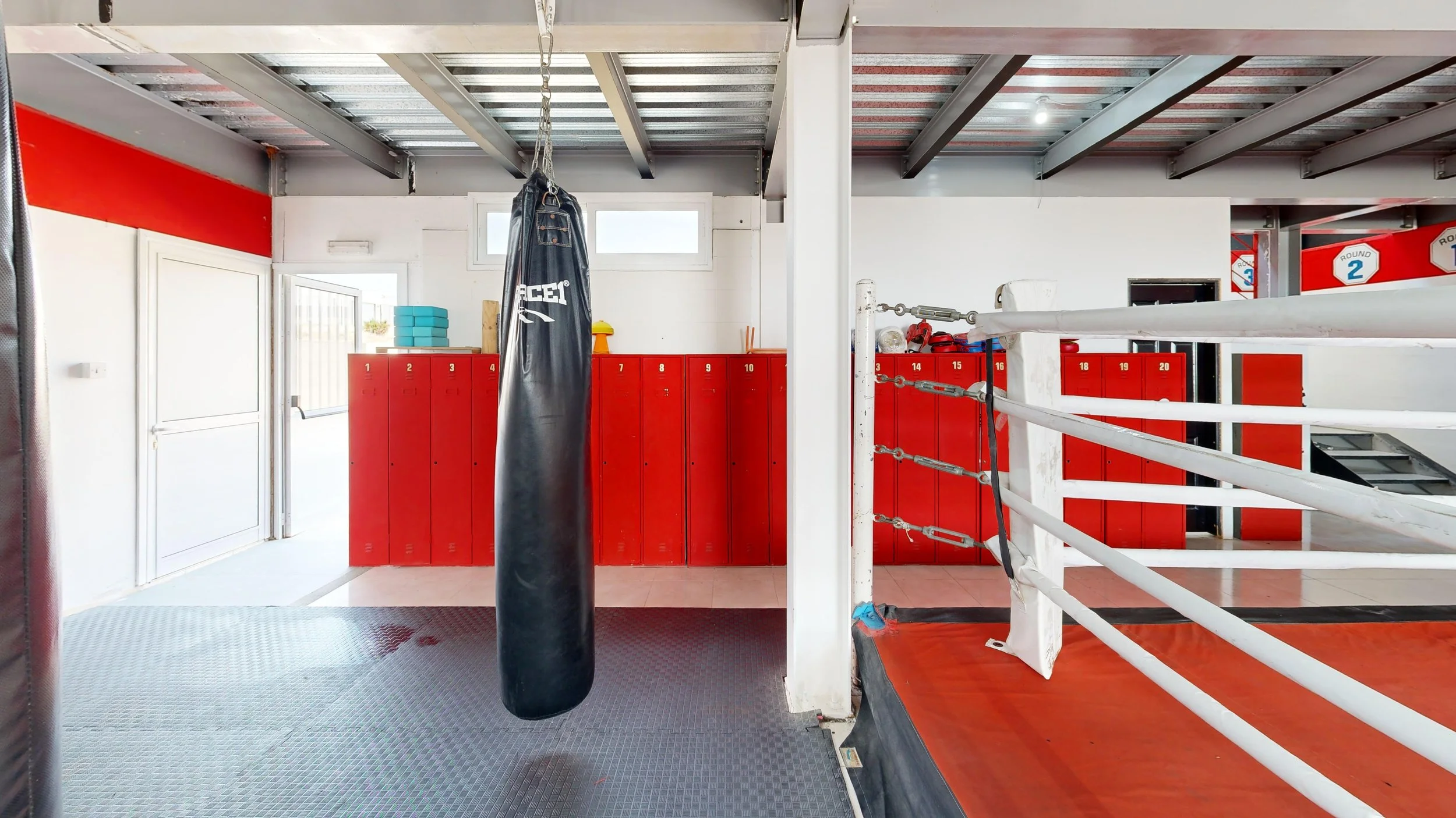 Boxing gym with red lockers, a punching bag hanging in front of lockers, and a boxing ring with white ropes and red padded floor.