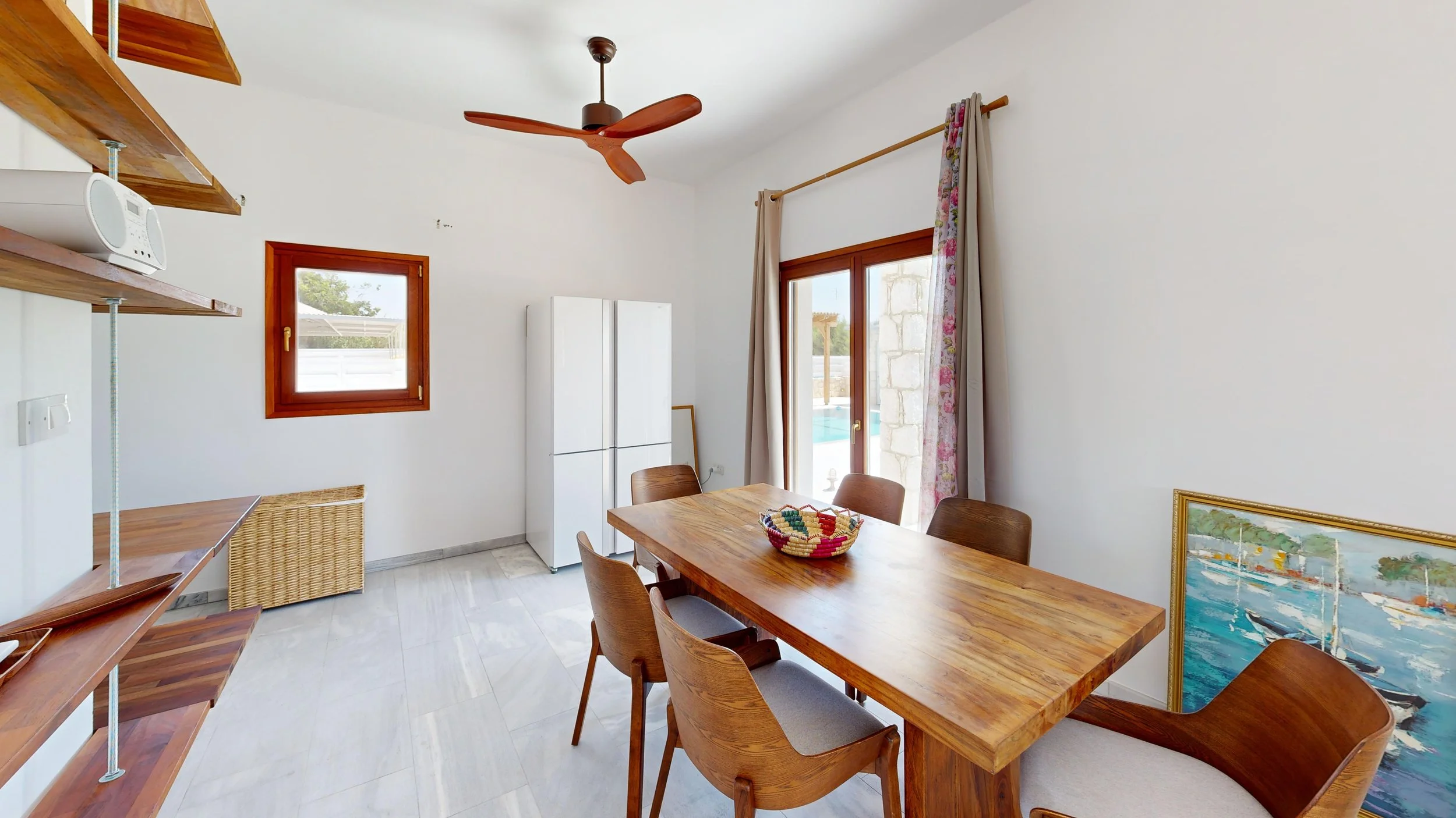 A dining room with a wooden table, six chairs, a white refrigerator, a framed picture on the floor, a small window, and a sliding glass door with curtains leading outside.