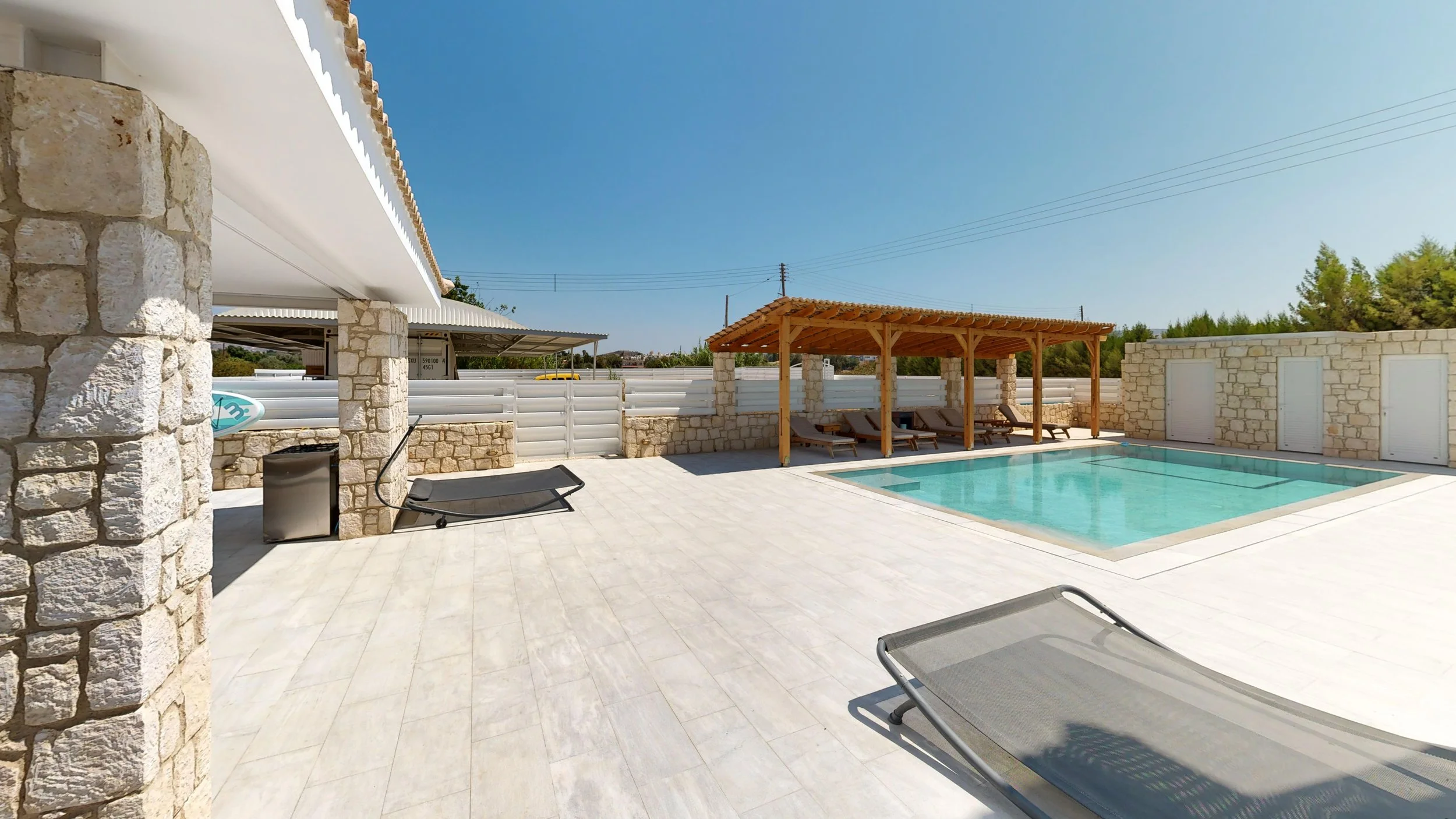 Exterior view of a swimming pool area with stone walls, a wooden shaded lounging area with chairs, and a clear blue sky.
