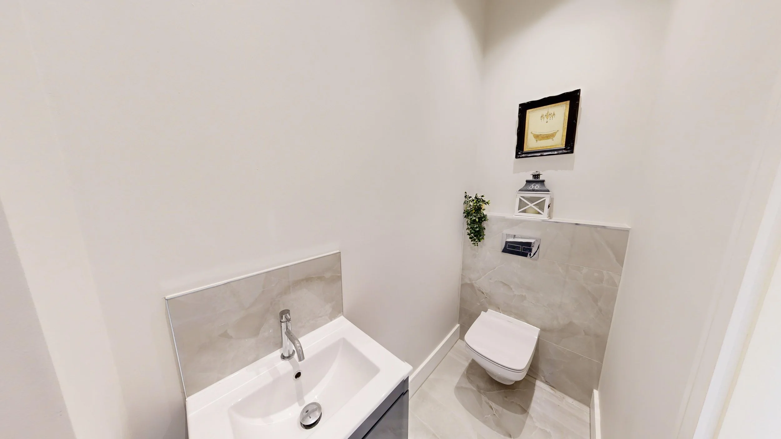 A small bathroom with a white sink, a wall-mounted toilet, and decorative items on a white ledge, including a framed picture, a lantern, and a small plant, with cream-colored tiled floor and white walls.
