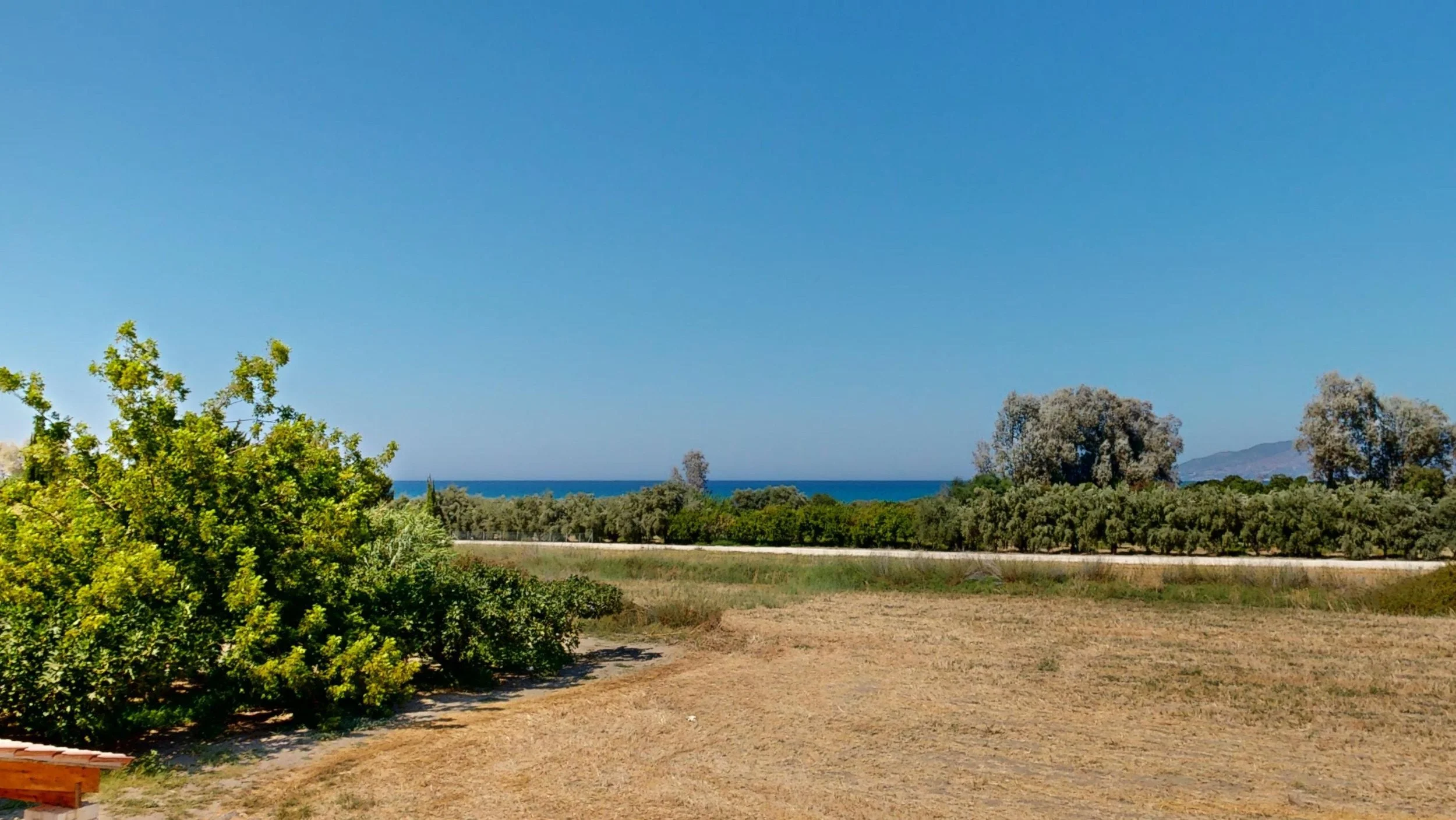 A landscape with green bushes and trees, a dirt field, a road in the middle distance, with the ocean and mountains in the background and a clear blue sky above.