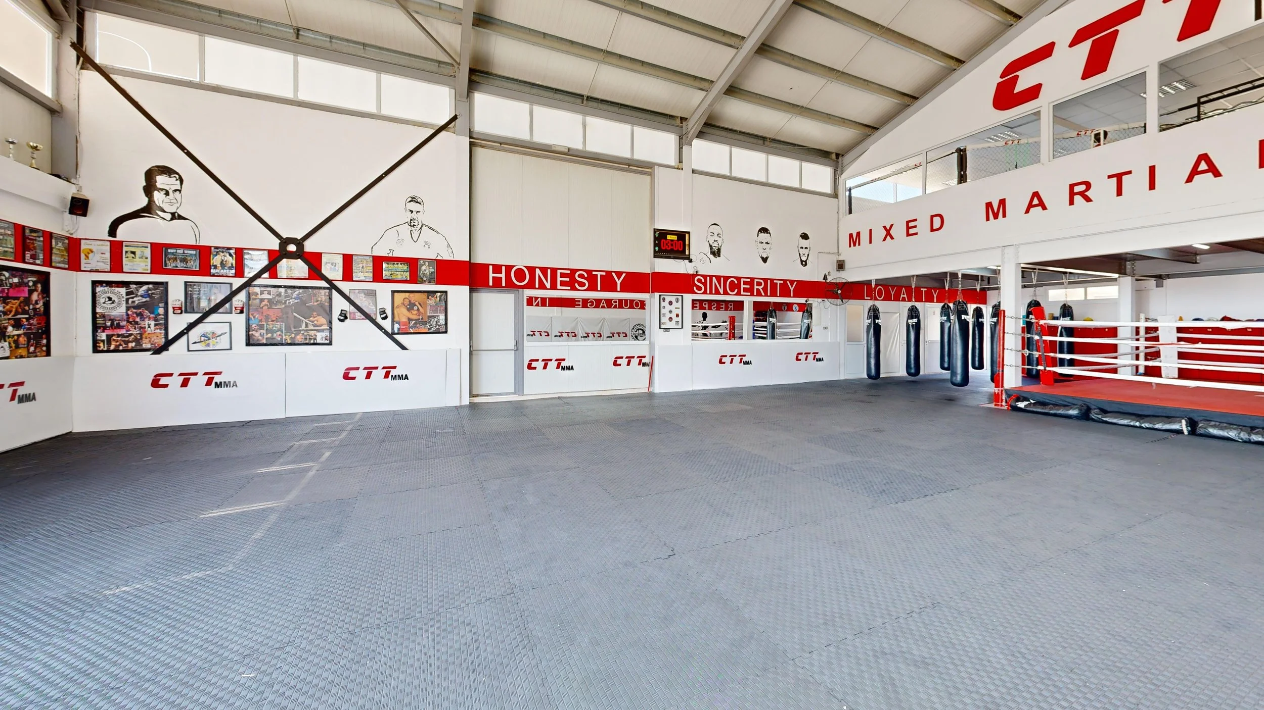 Inside a martial arts gym with punching bags, a boxing ring, and wall art including portraits and motivational words like honesty, sincerity, and loyalty.