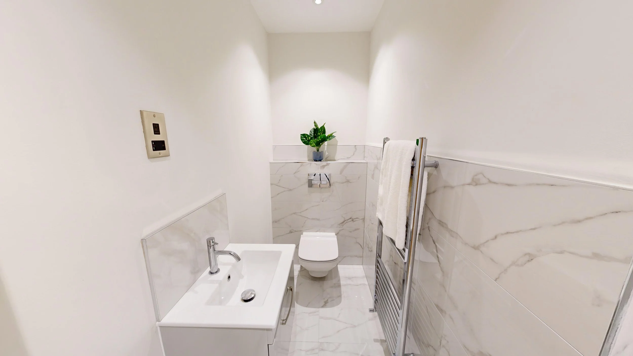 Small modern bathroom with white walls and marble tile accents. Contains a white toilet, a small white sink with a chrome faucet, a towel rack with a white towel, a potted plant on a marble ledge, and a wall-mounted control panel.
