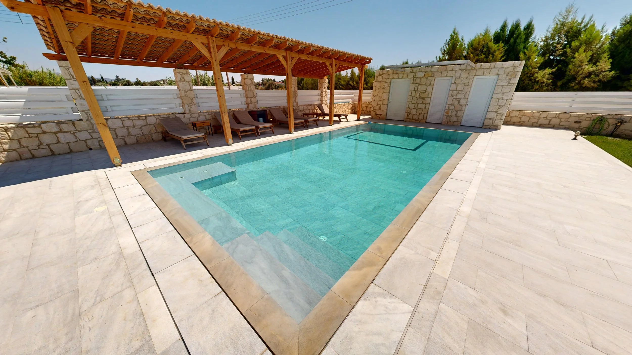A private swimming pool with a clear blue color, surrounded by light-colored tiled deck, and a shaded area with wooden lounge chairs and a small side table. The background includes a stone wall with three doors, a row of trees, and a bright clear sky.