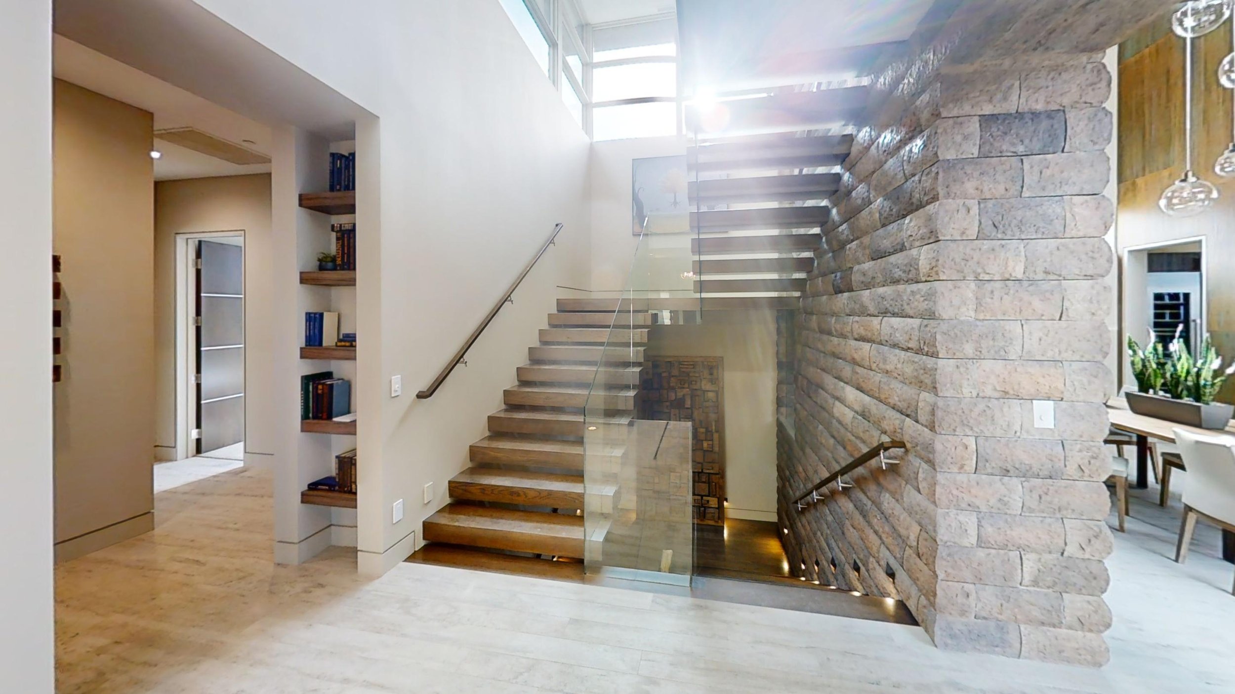 Interior view of a modern home with a staircase featuring wooden steps, glass railing, and a stone accent wall. Sunlight streams through upper windows, illuminating the space.