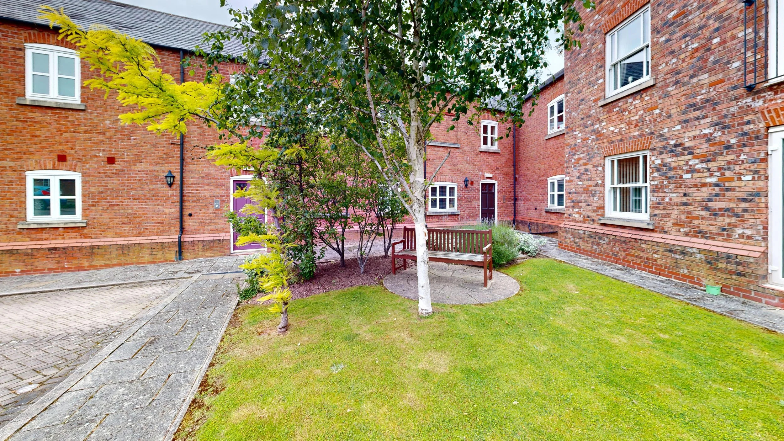 Courtyard with green grass, trees, and benches surrounded by red brick buildings with white trim on windows and purple doors.