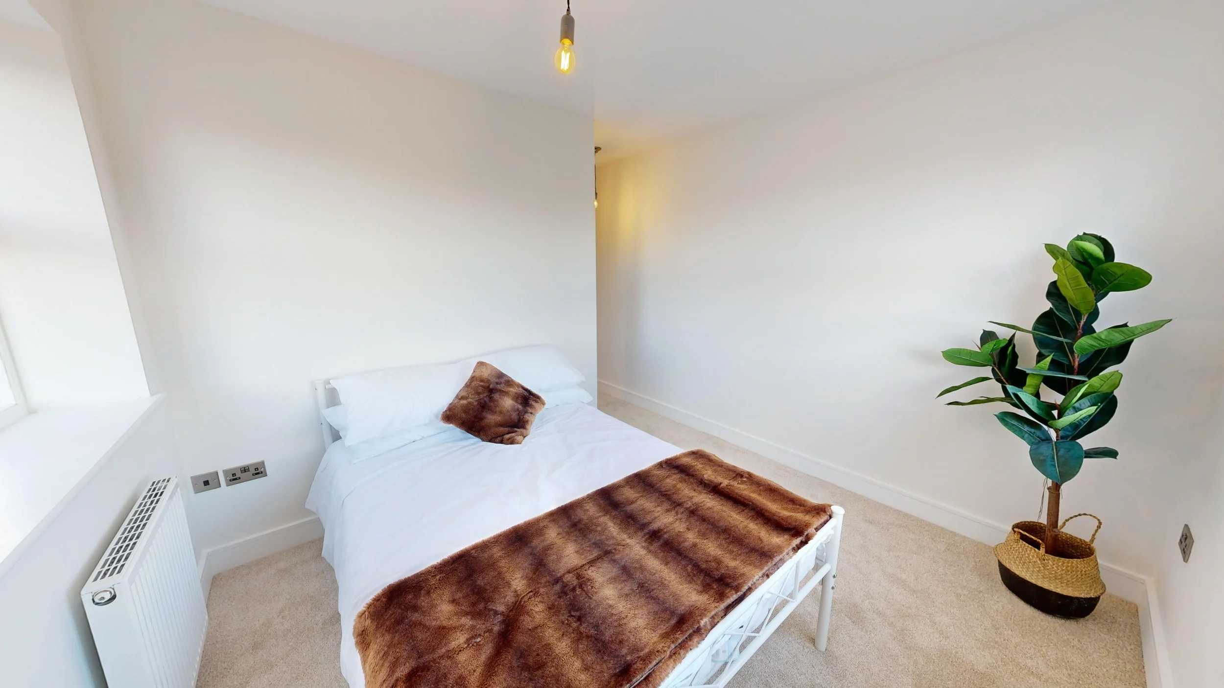 A minimalist bedroom with a white bed, brown fur blanket, matching brown fur pillow, beige carpet, a large window on the left, a potted plant in a black and beige woven basket on the right, and an exposed lightbulb hanging from the ceiling.