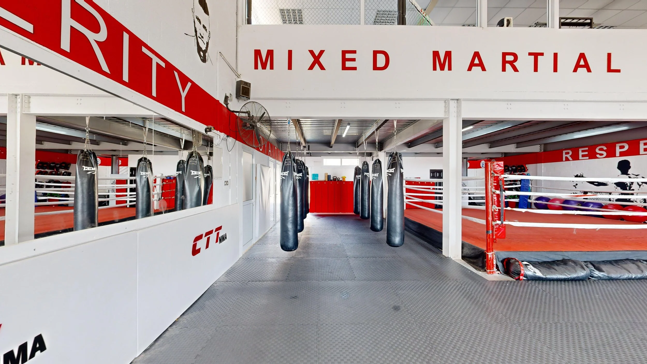 Interior of a mixed martial arts gym with punching bags hanging in the center, a boxing ring to the right, and training equipment on the walls, with signs that say 'Mixed Martial' and 'Respect' in bold red letters.