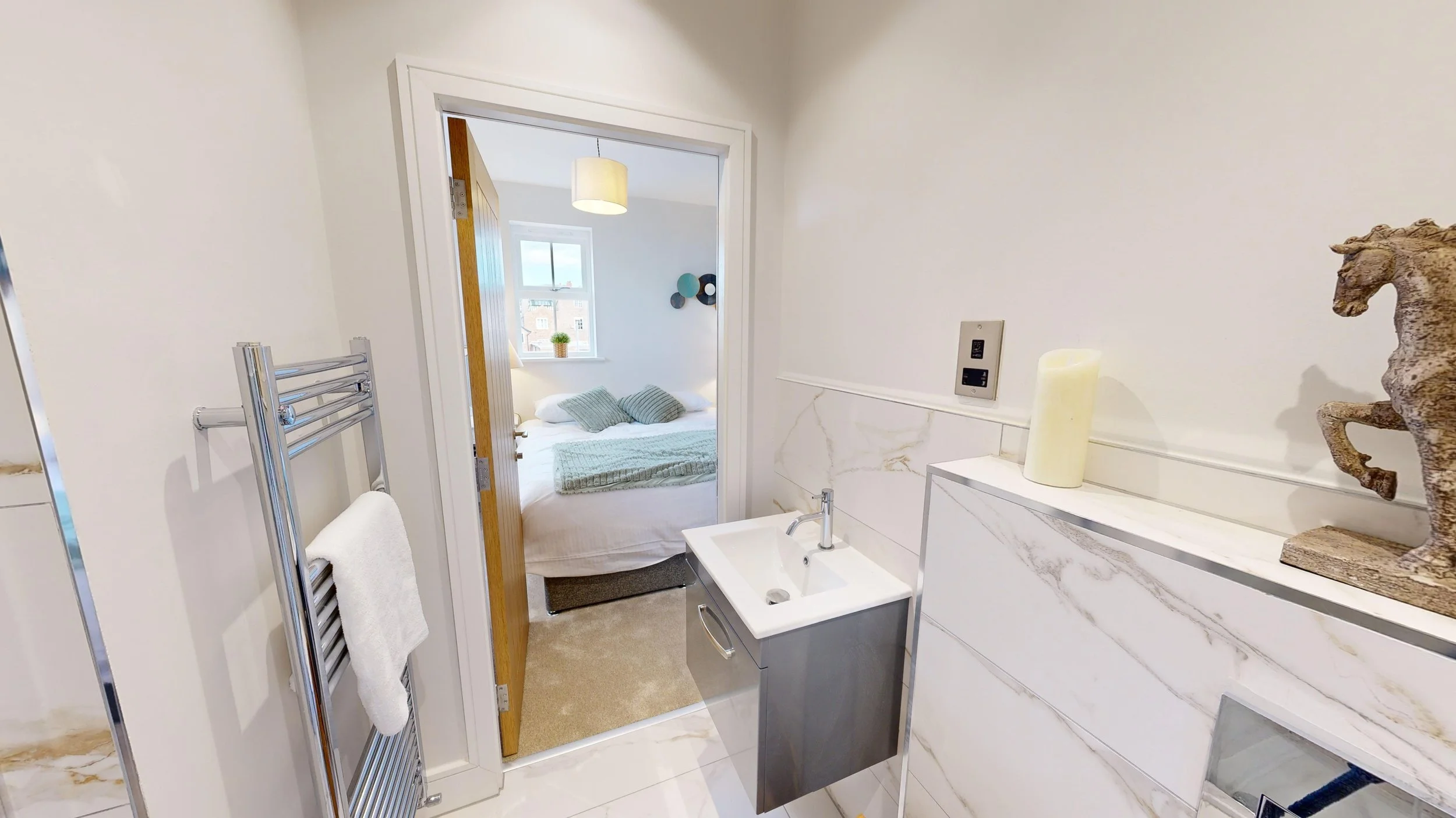 View of a small bathroom with a wall-mounted sink, a towel rail with a white towel, decorative items on a shelf, and an open door leading to a bedroom with a bed, pillows, a window, and wall decor.
