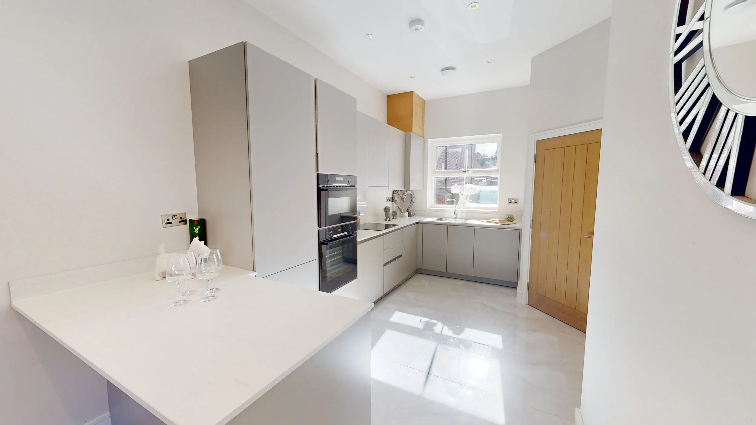 Modern white kitchen with light wood accents, a window overlooking outdoors, and three wine glasses on a white countertop.