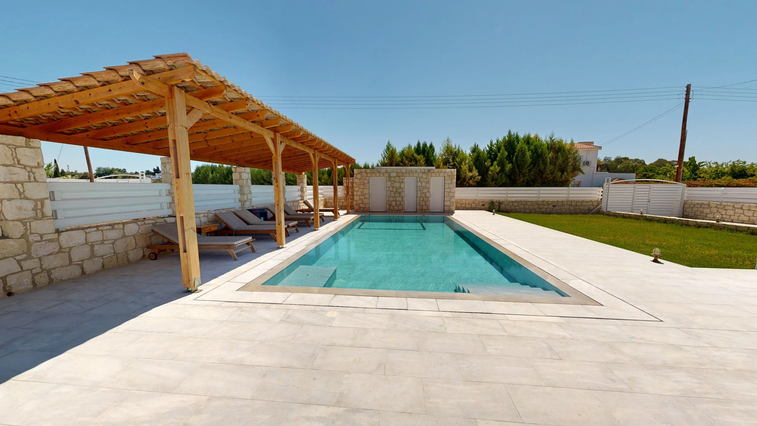 A rectangular swimming pool with steps and a spa, surrounded by white stone tiling and a grassy yard. There is a shaded area with lounge chairs under a wooden canopy with a tiled roof. The background shows a stone wall, trees, and a clear blue sky.