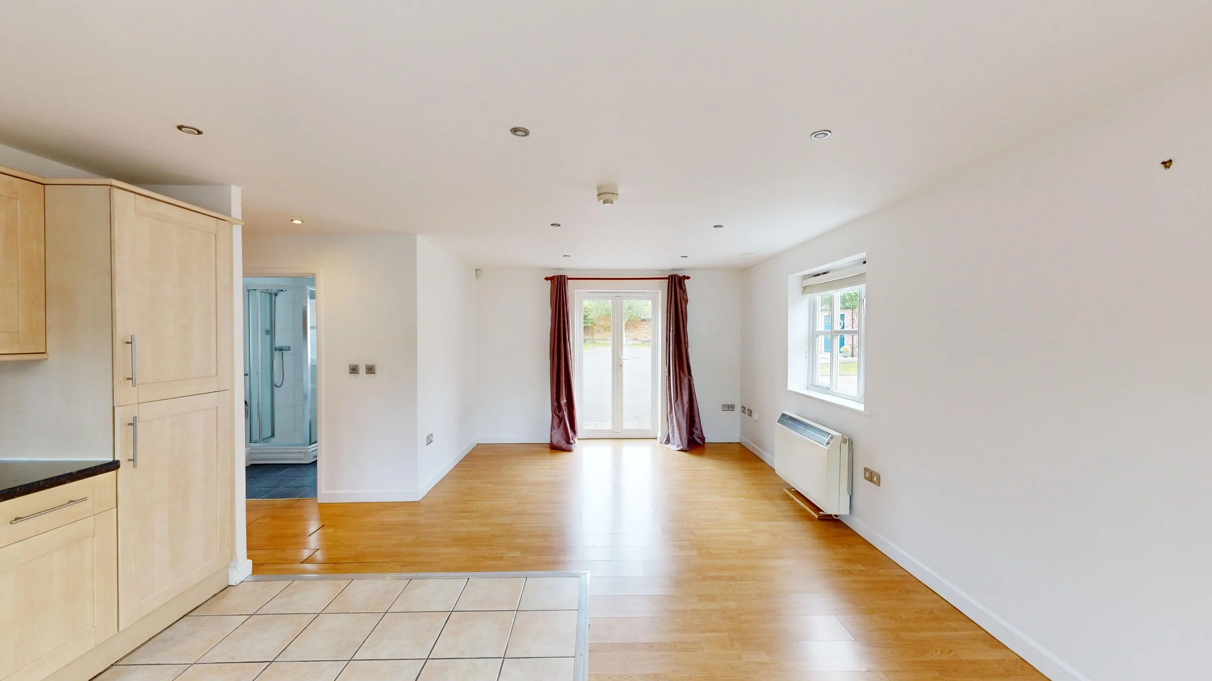 Empty living room with large window, glass door, hardwood floor, and kitchen area with tiled floor on the left. Curtain panels are hanging on the glass door, a radiator is below the window, and a corner shower stall is visible through an open doorway.