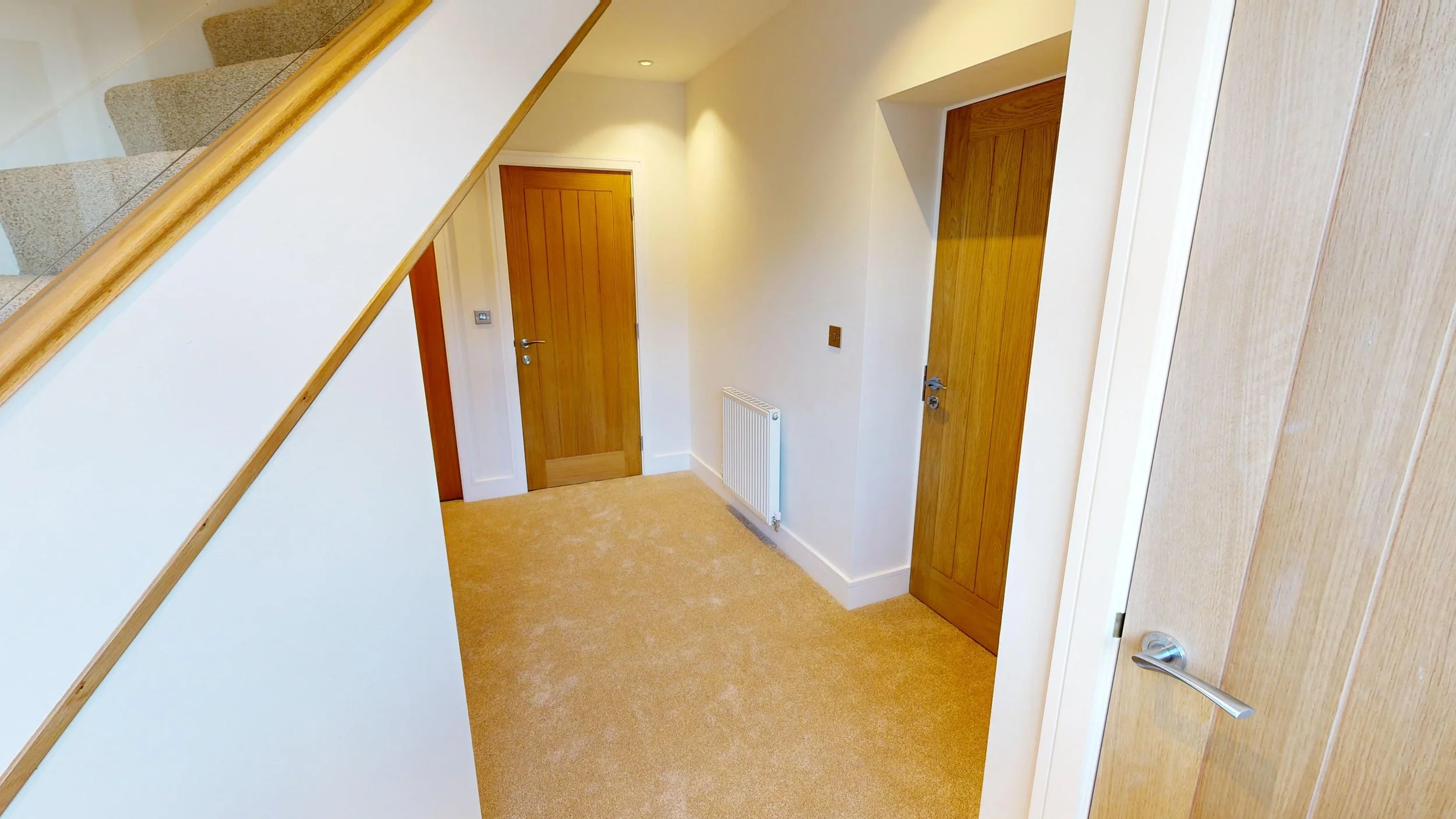 Interior view of a hallway with beige carpet, white walls, and wooden doors. A staircase is partly visible on the left side of the image.