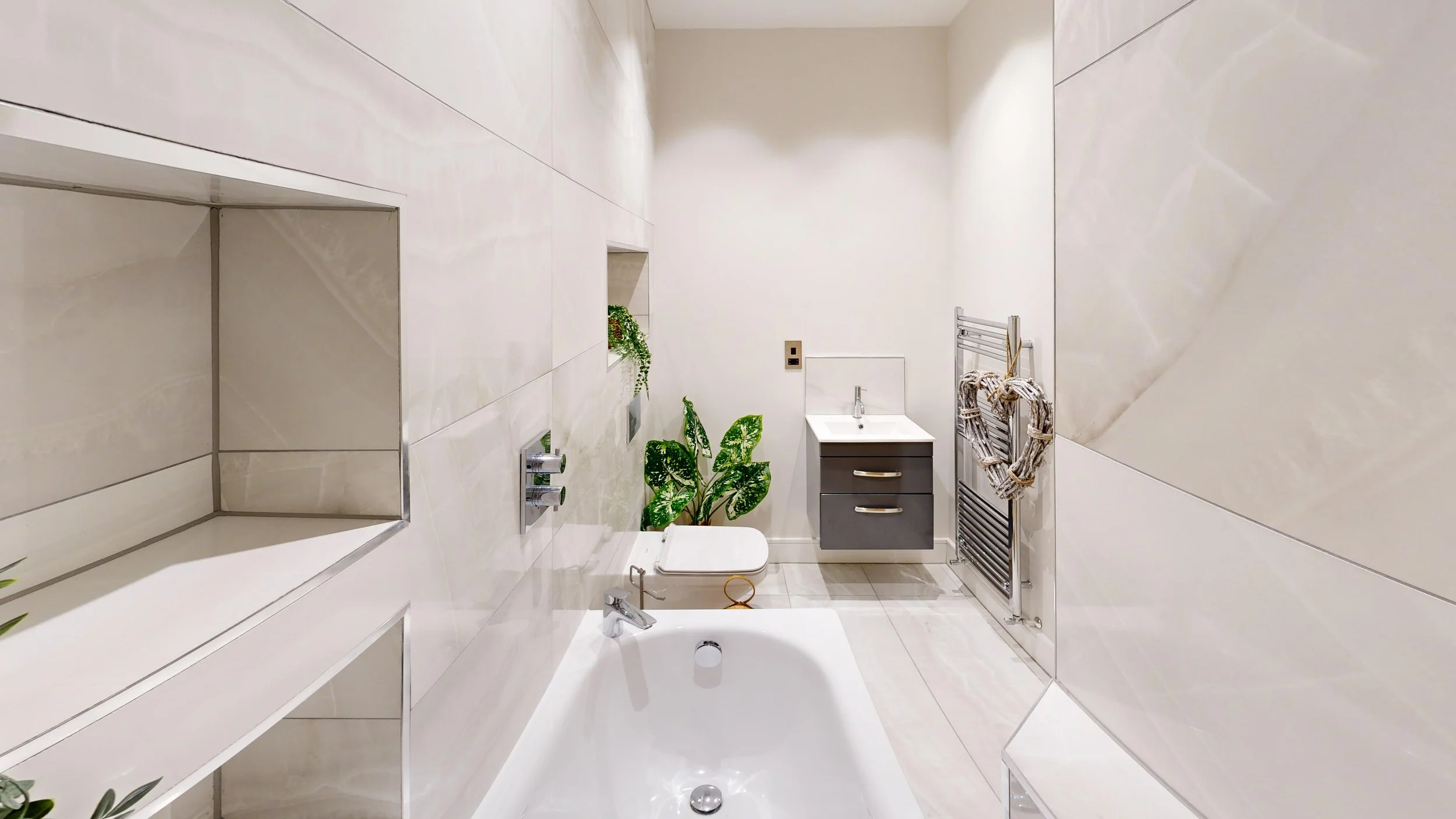 Modern bathroom with a bathtub, a small sink with gray cabinet, plants, towel rack, and minimal decor, all in white and gray tones.