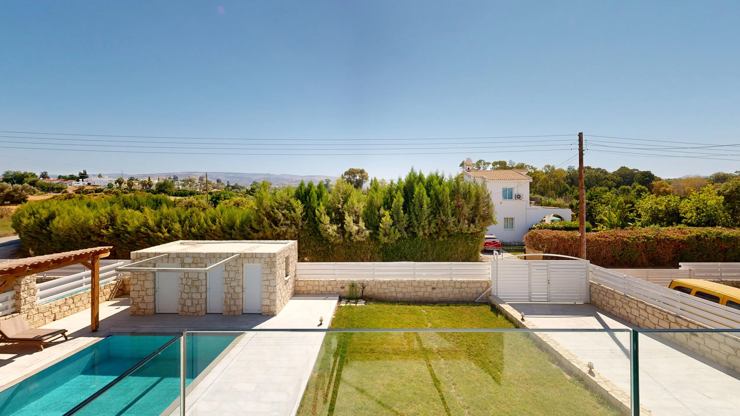 Residential backyard with swimming pool, stone shed, lawn, white fence, and surrounding trees under clear blue sky.
