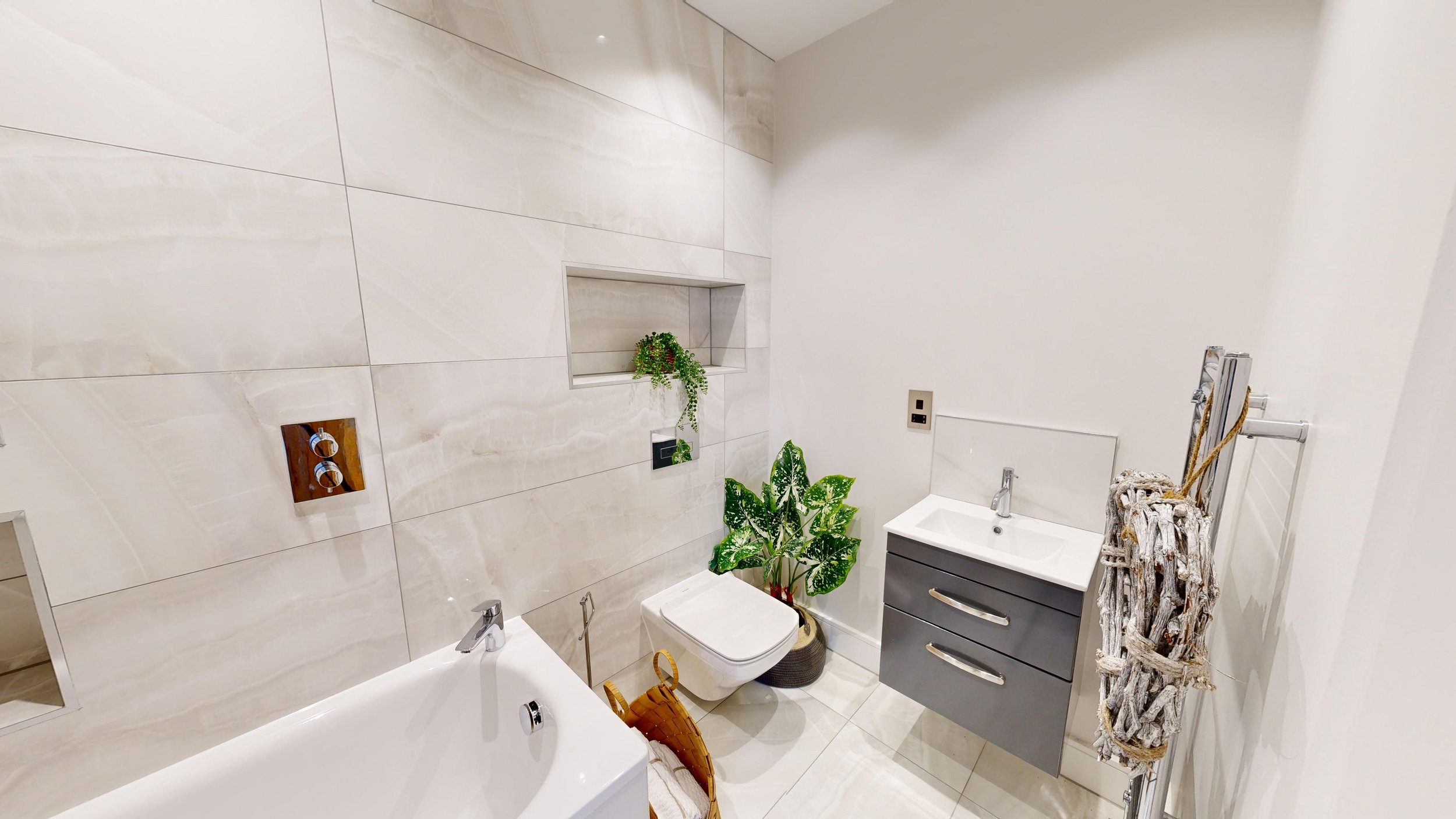 Modern bathroom with white marble walls, a bathtub, a wall-mounted toilet, a gray vanity with a white sink, plants, and a towel rack with decorative towels.