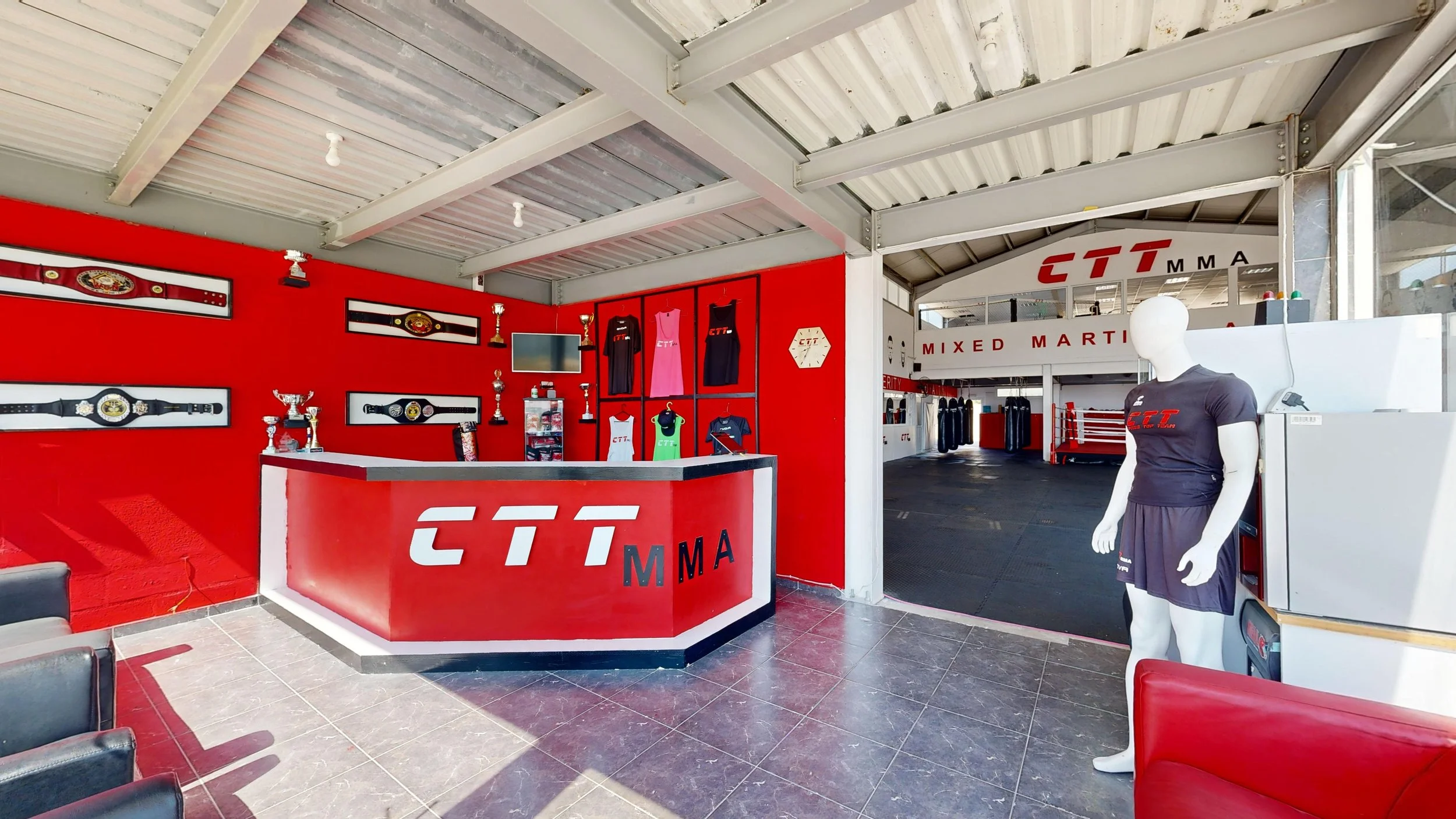 Interior of a martial arts gym with a red reception desk featuring the CTT MMA logo, trophies on display, martial arts apparel on the wall, and a mannequin dressed in black MMA gear outside the entrance.