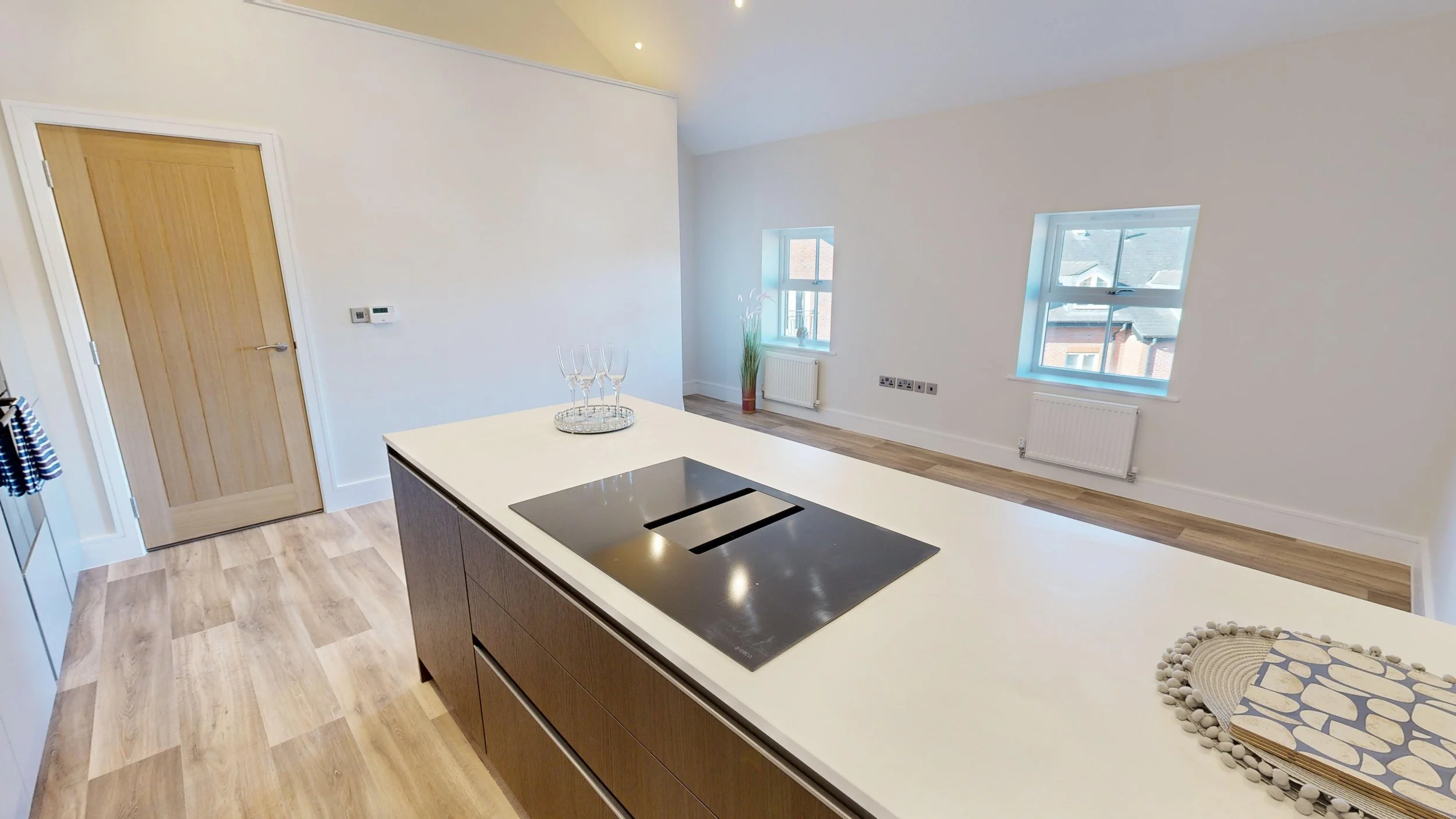 Modern kitchen with white island countertop, electric cooktop, and wooden cabinetry. Open living space with two windows, hardwood floors, and minimal decor.