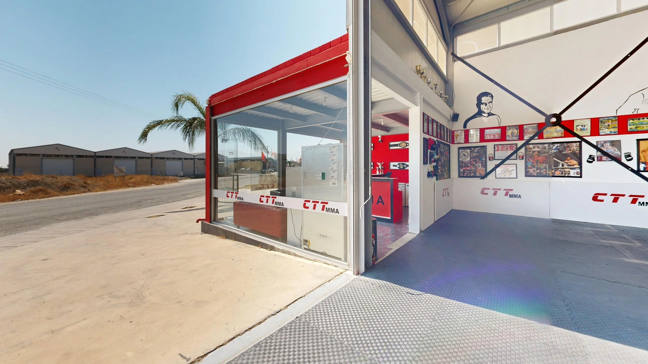View of a small boxing gym with a glass front, red and white decor, and pictures on the wall, located on a street with warehouses and palm trees in the background.