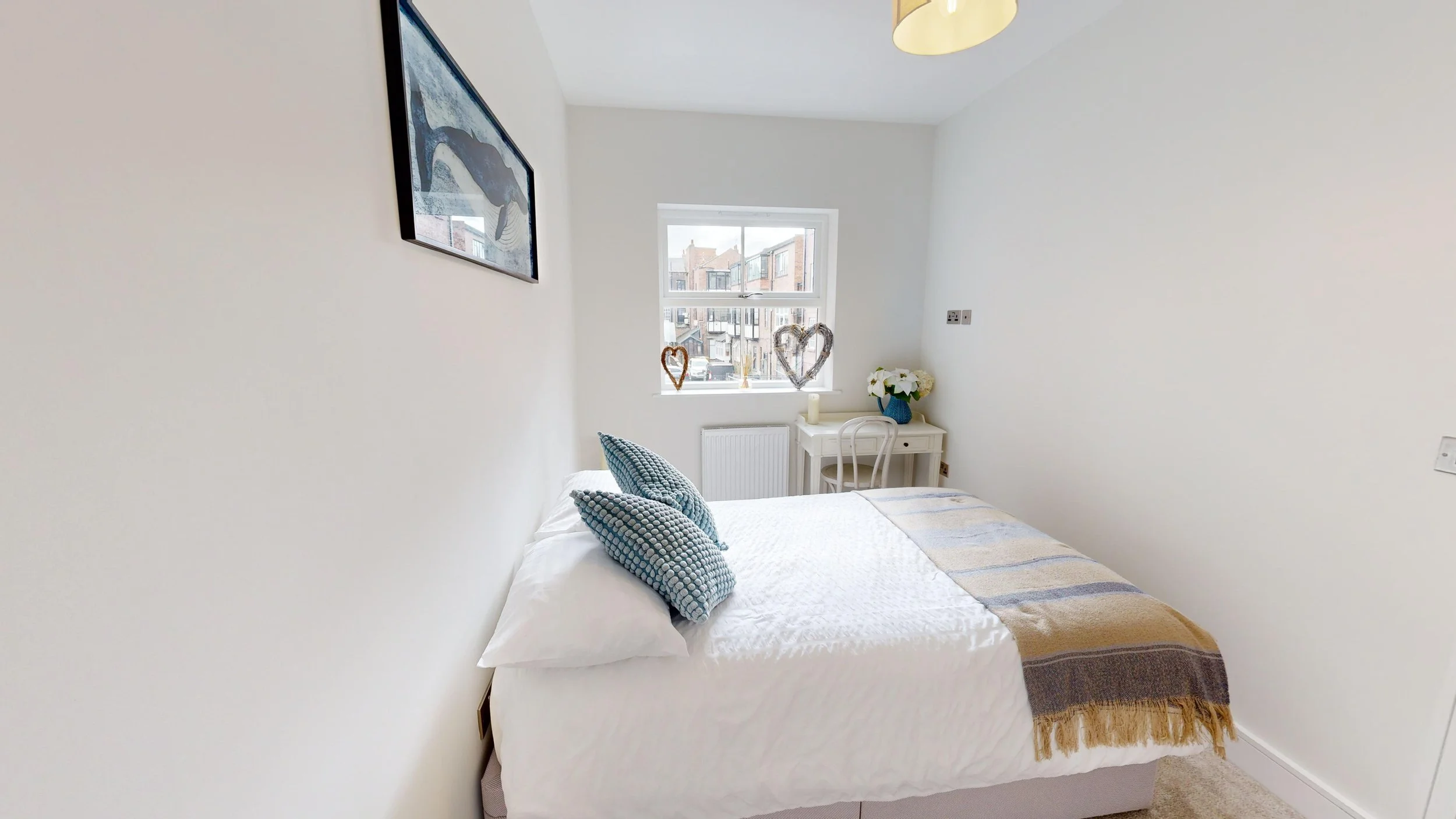 A bright bedroom with a white bed, blue patterned pillows, a colorful blanket, a small white desk with a white chair, a vase of white flowers, and two heart-shaped decorations on the window ledge, with a window showing neighboring buildings.