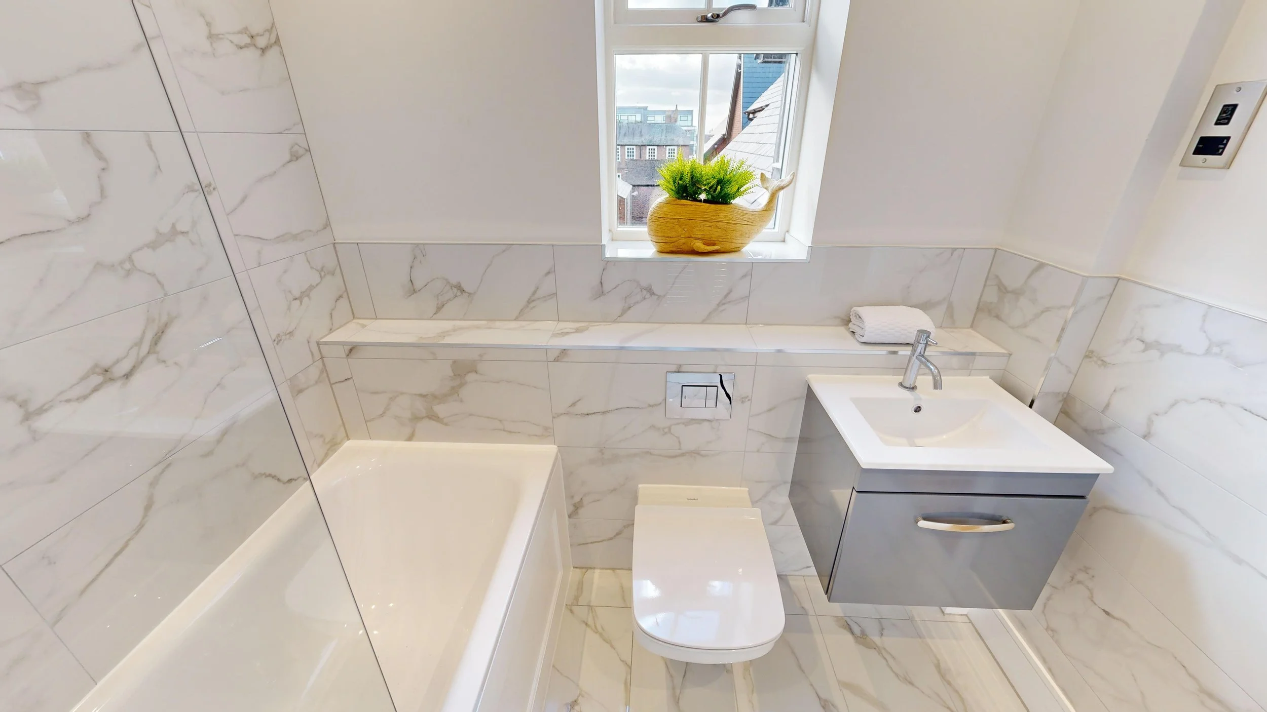 Small modern bathroom with white marble walls and floor, a window with a potted plant, and fixtures including a toilet, sink, and bathtub.