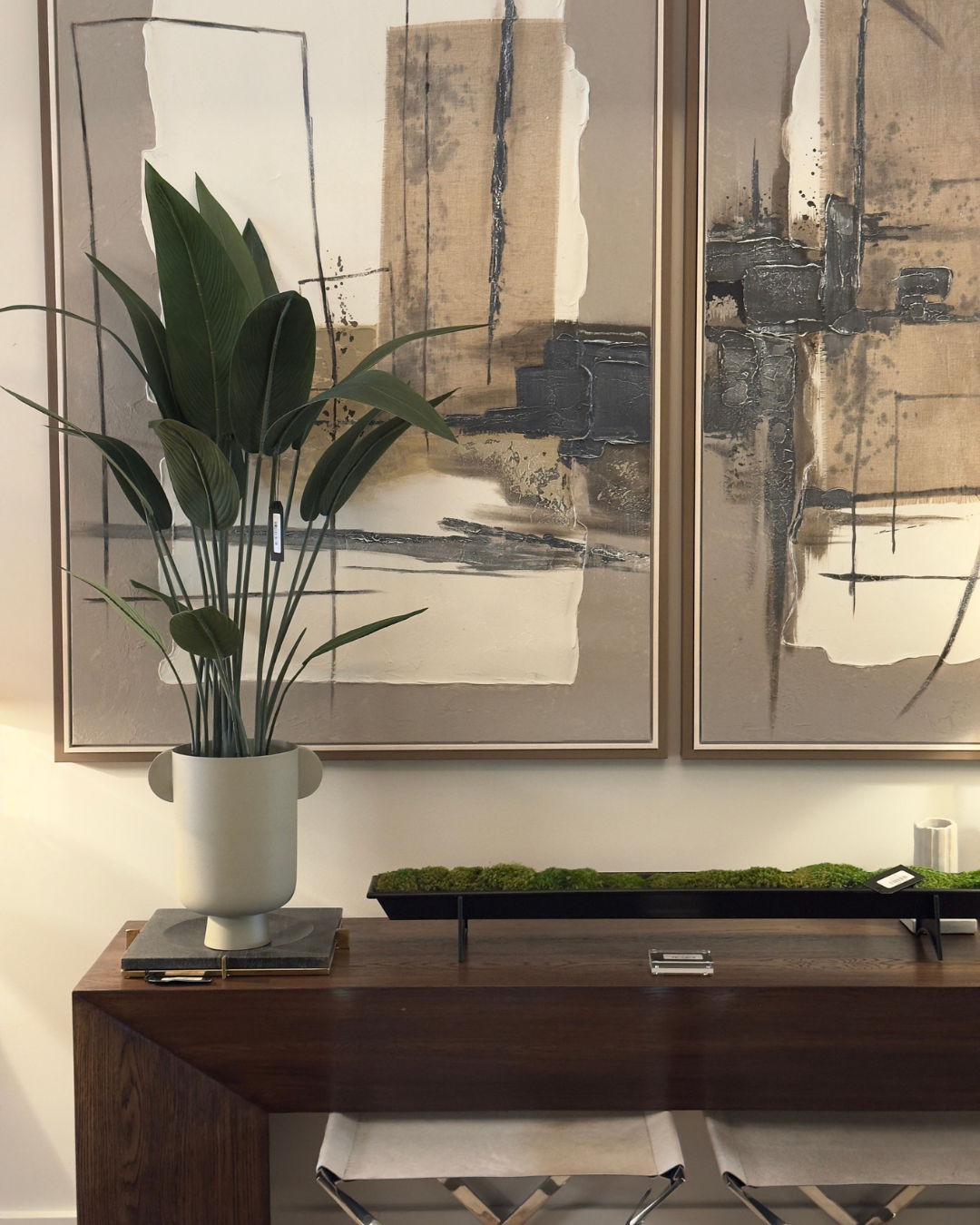 Interior decor with large abstract paintings, a tall green plant in a white vase on a wooden console table, with moss and other decorative objects, and two beige cushioned stools in front. Modern furniture and home decor store in New Orleans.