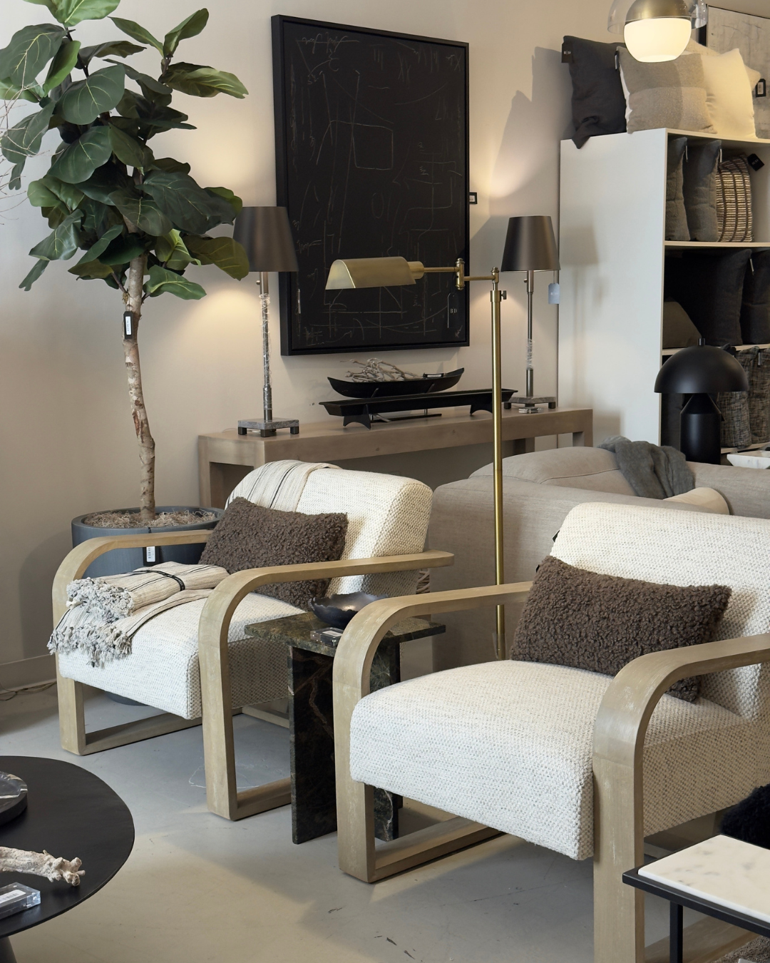 Modern furniture and home decor store in New Orleans. A modern living room with two beige armchairs featuring light wood frames, brown textured pillows, a black marble side table, a sofa, stylish lamps, a large potted plant, and wall art.