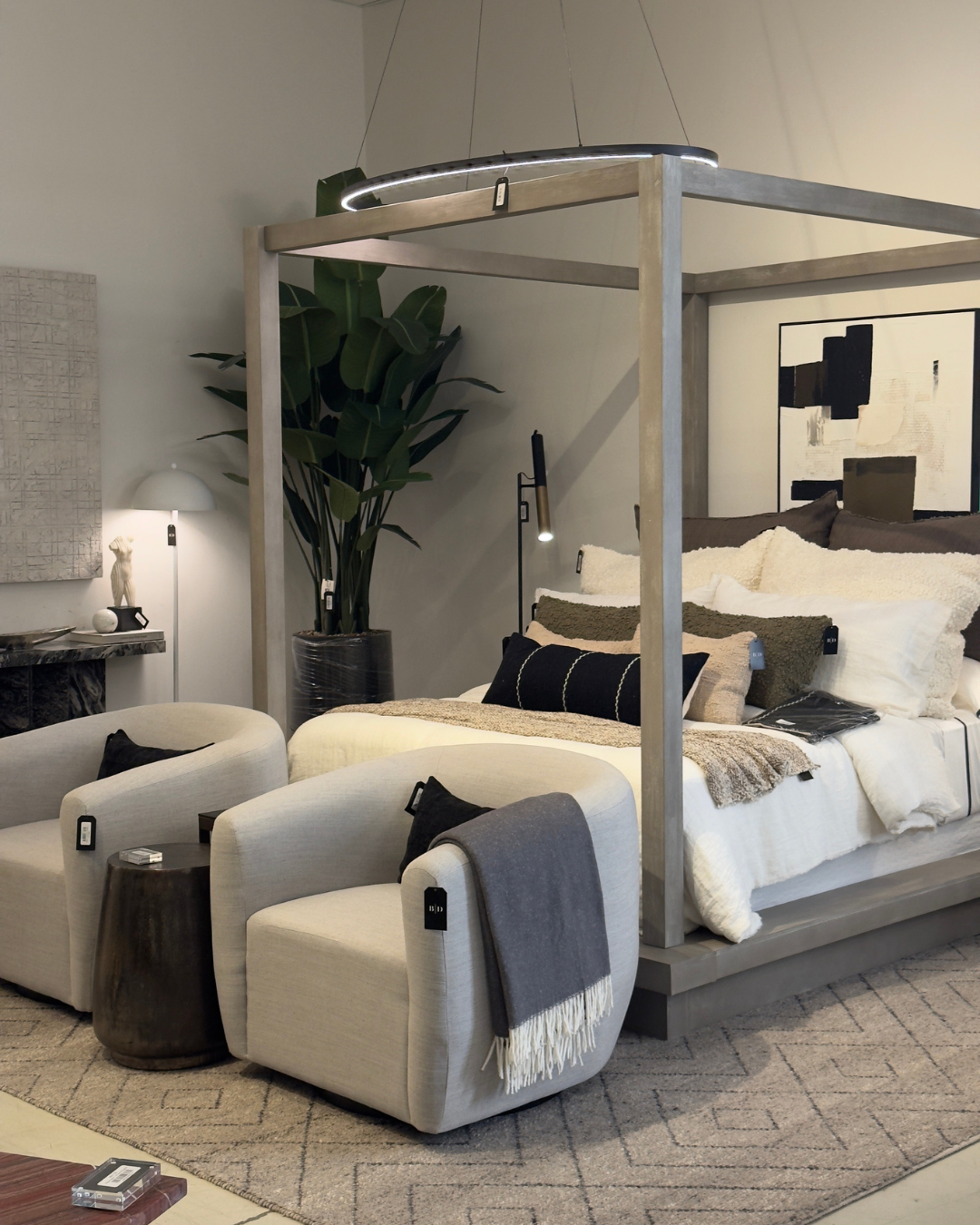 A modern bedroom with a four-poster bed, beige armchairs, a large potted plant, and abstract artwork on the wall. Modern furniture and home decor store in New Orleans.