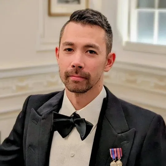 A man with short dark hair and a beard, wearing a tuxedo with a bow tie and medals, sitting indoors.