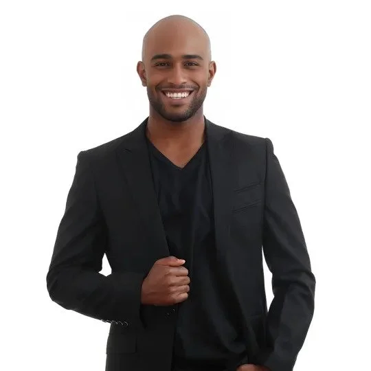 A man with a shaved head and beard smiling, dressed in a black blazer and shirt, standing against a white background.