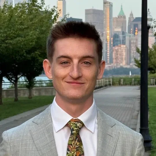 Young man in a light gray suit and floral tie standing outdoors with city skyline in the background.