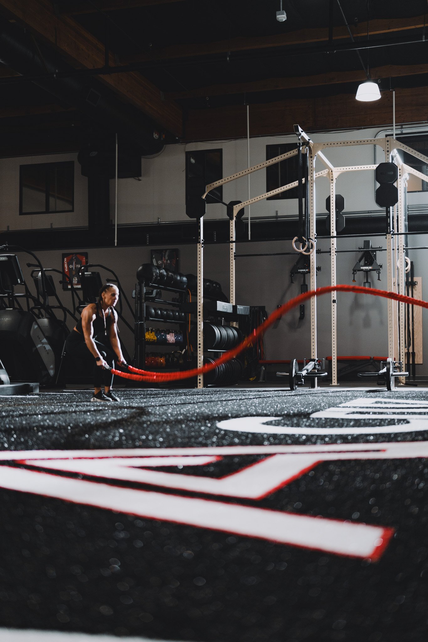 A woman performing battle ropes workout in a gym with exercise equipment and a black and red mat on the floor.