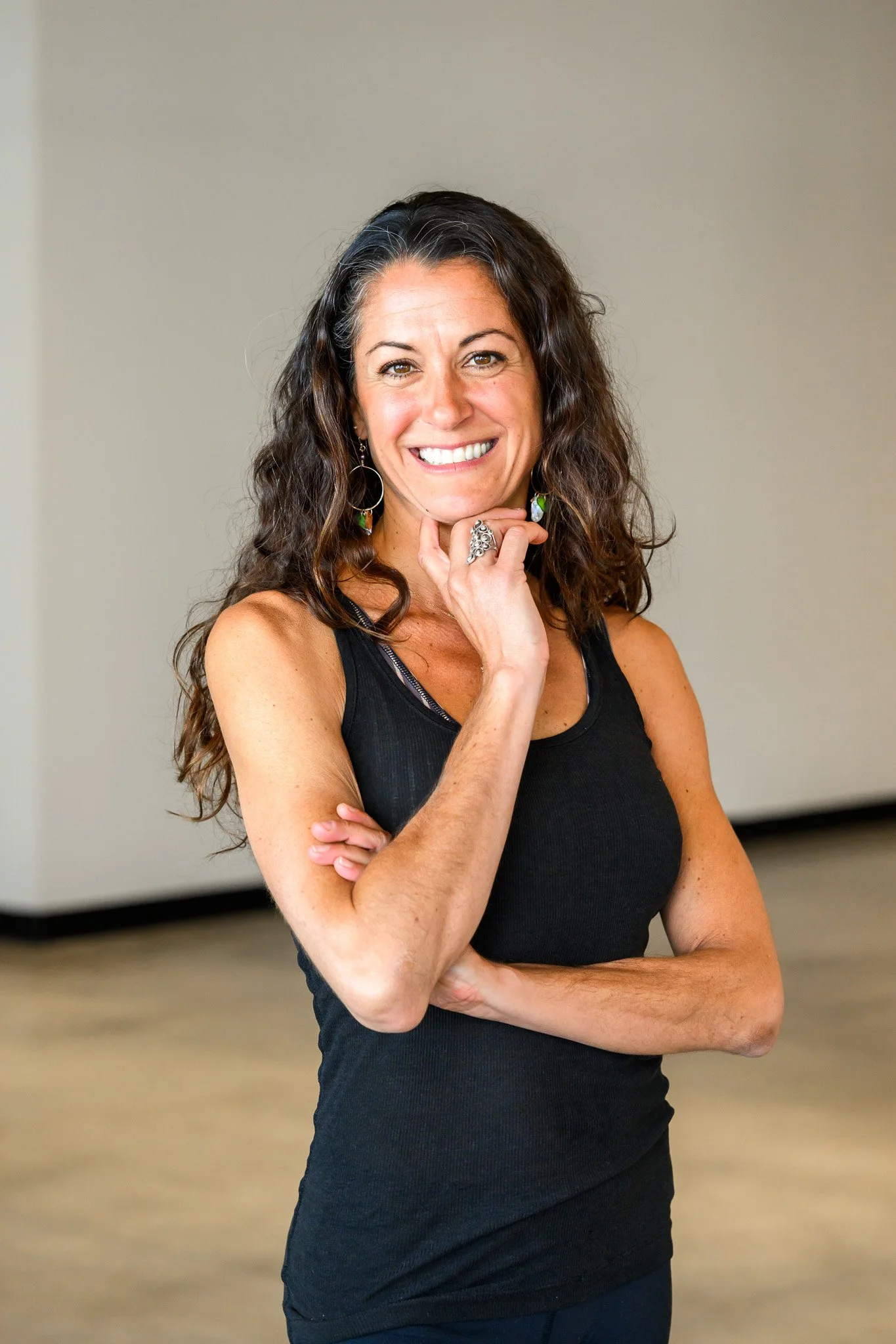 Woman with long curly hair smiling and posing in a sleeveless black top, wearing hoop earrings, in a neutral indoor setting.