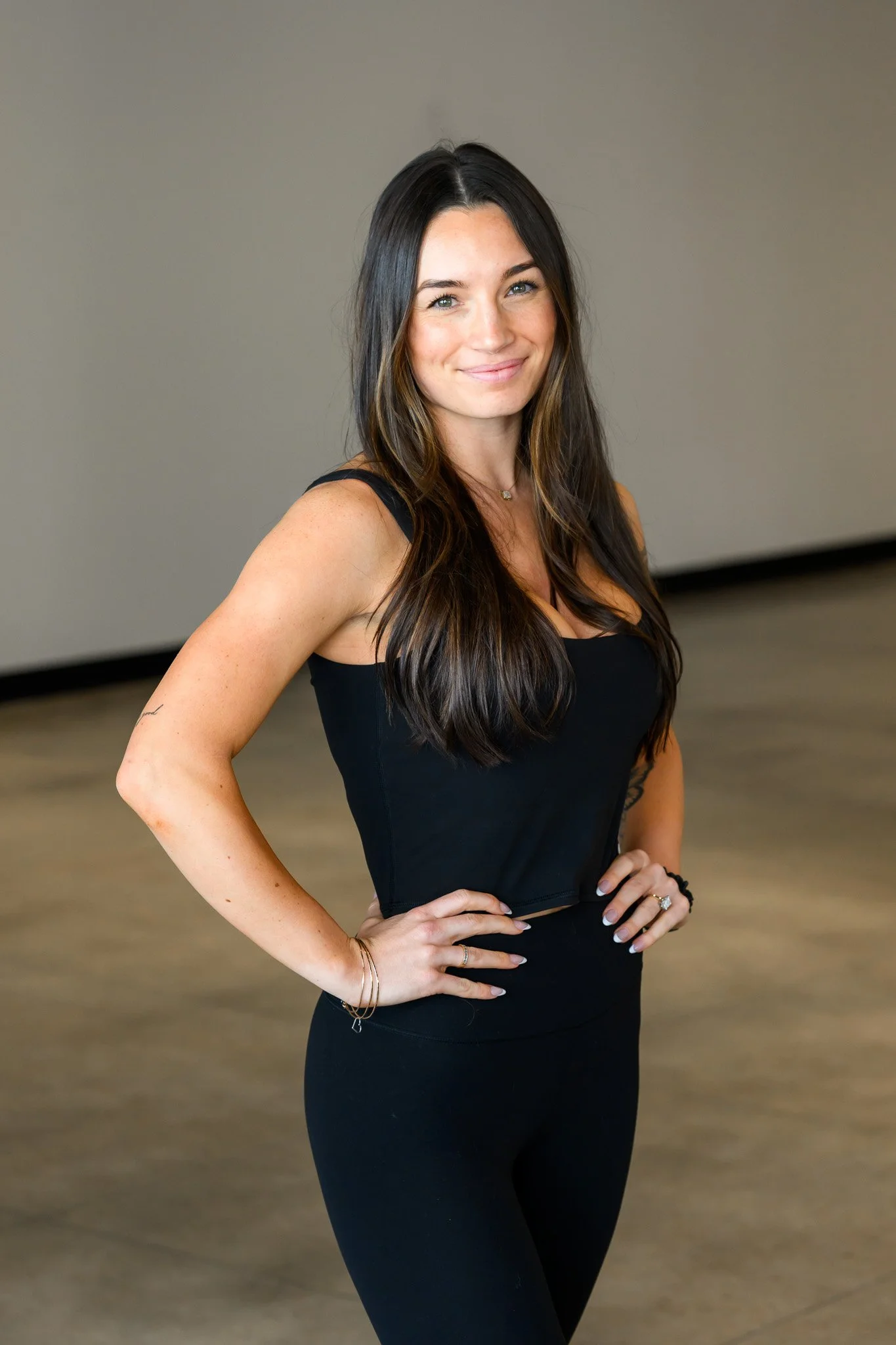 Smiling woman in a black tank top and leggings with long brown hair standing indoors.
