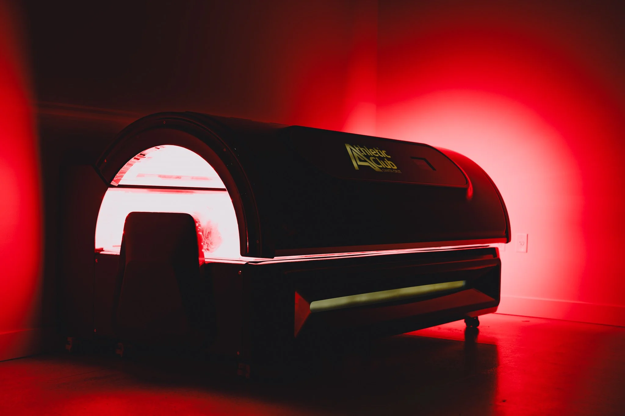 Red light therapy bed with a digital yellow logo that reads 'Athletic Club' on a wall with red lighting.