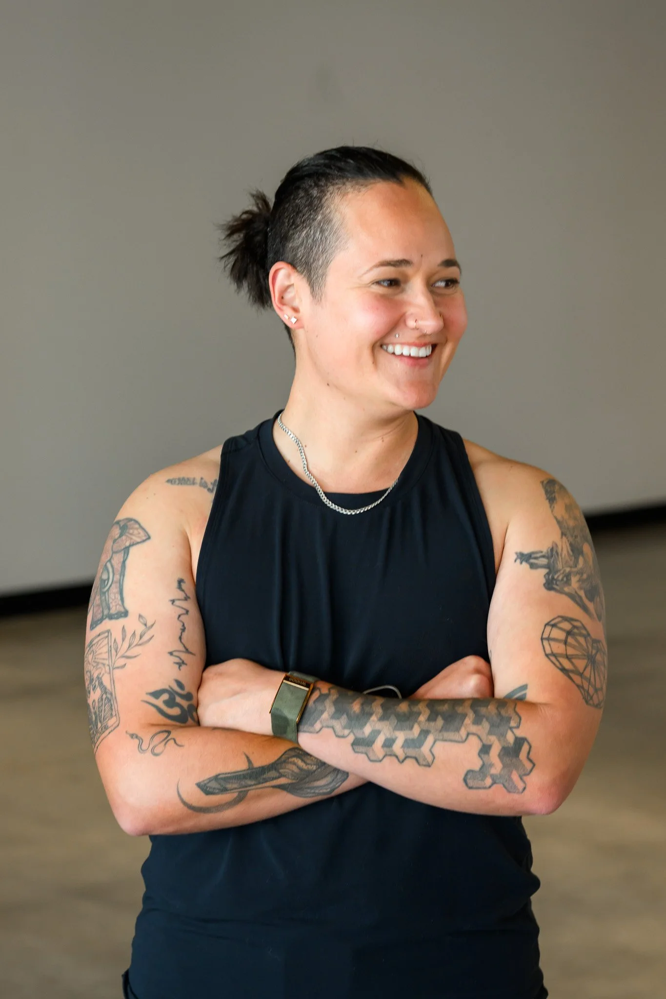 A woman with dark hair in a bun, wearing a black sleeveless top, smiling with arms crossed, showing multiple tattoos on her arms.