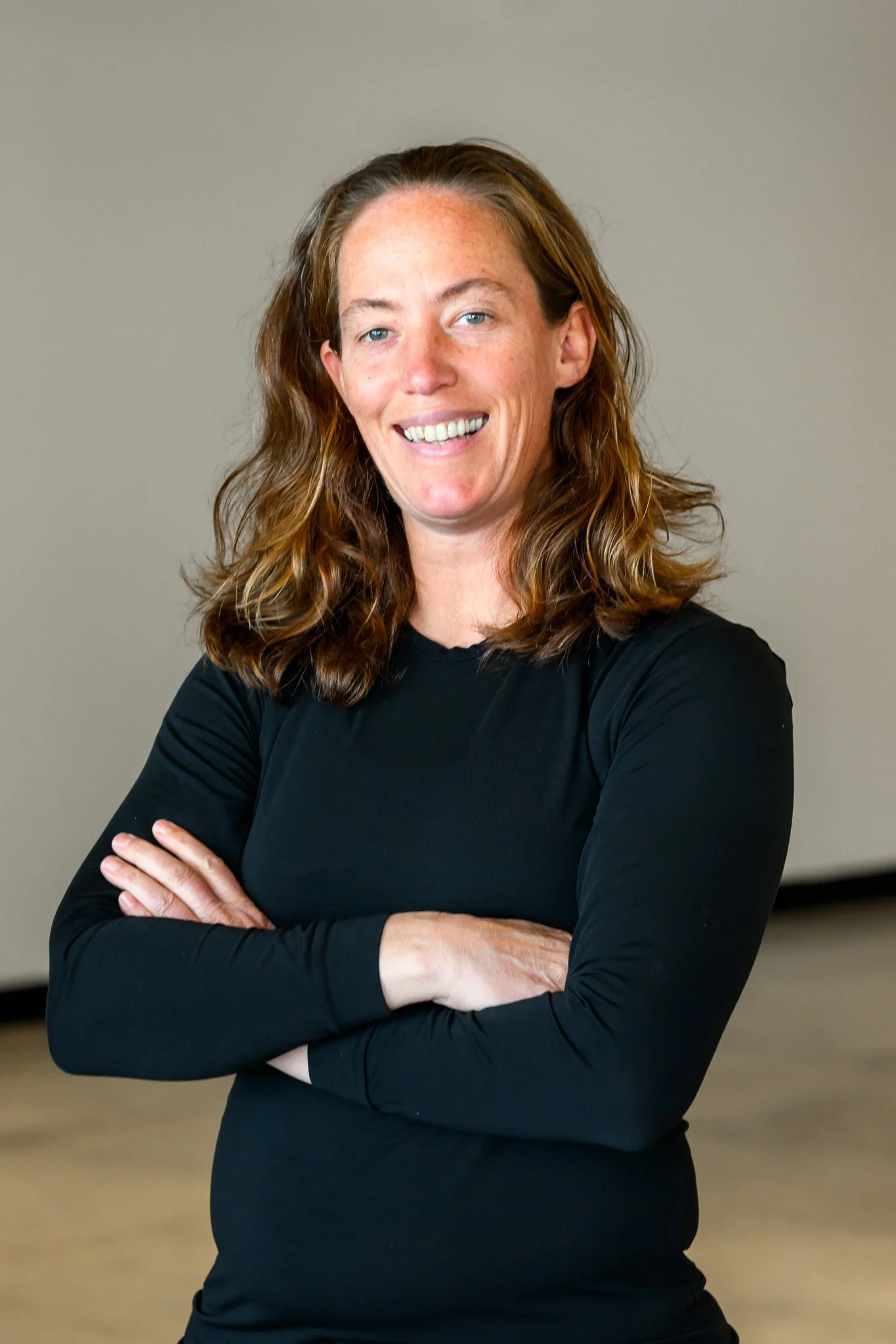 A woman with long, wavy, reddish-brown hair, smiling and crossing her arms, wearing a black long-sleeve top against a neutral background.