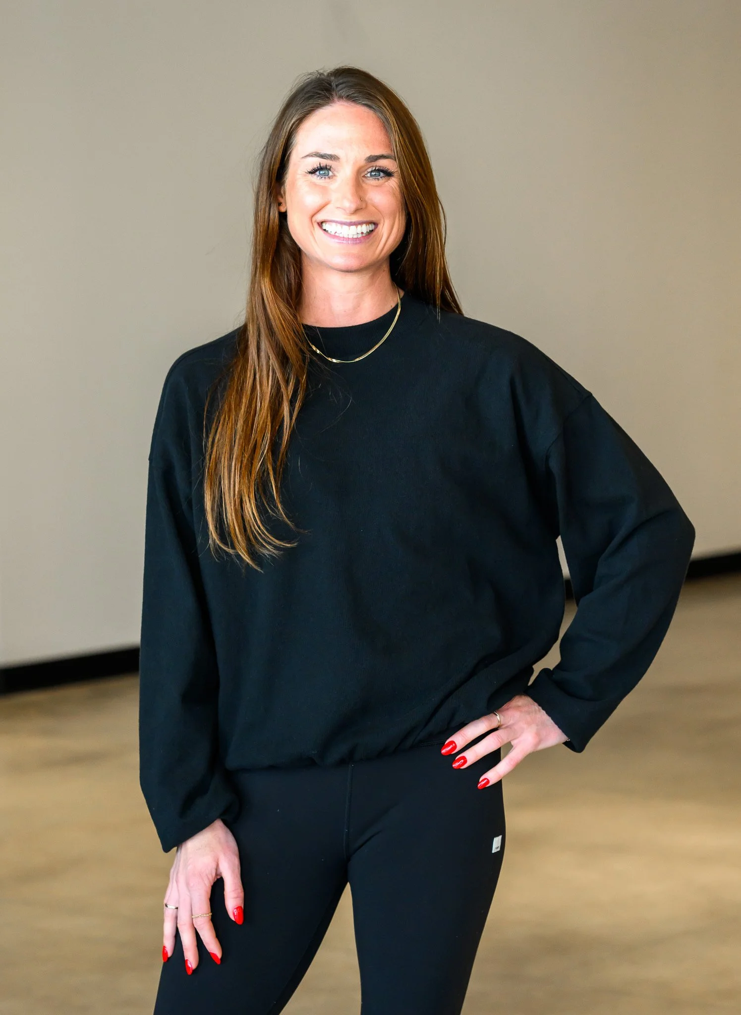 Woman with long brown hair, wearing a black long-sleeve shirt and black pants, smiling and standing with one hand on her hip in an indoor setting.