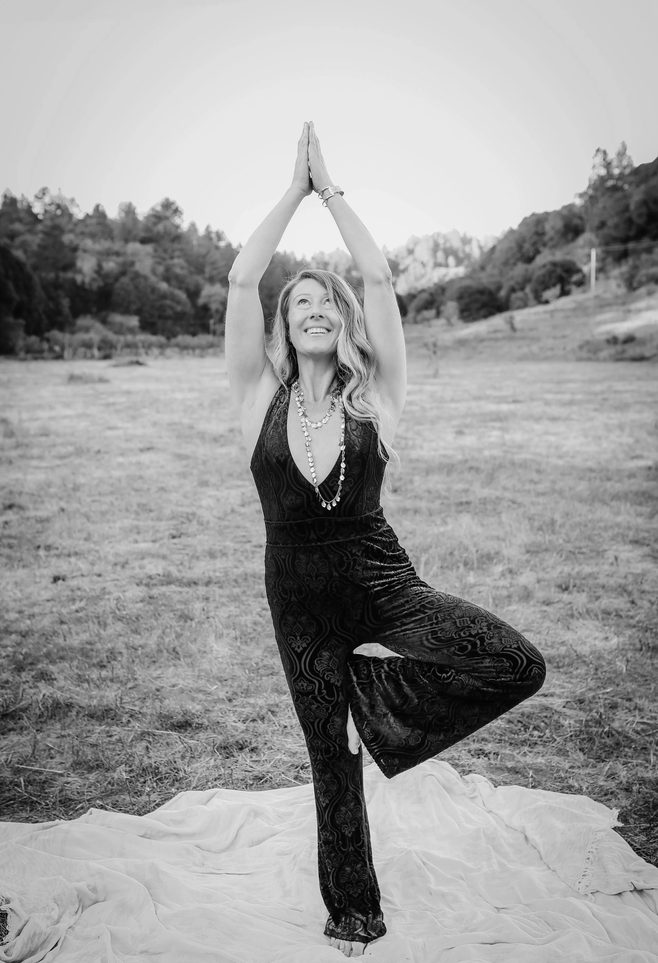 A woman practicing yoga outdoors on a blanket in a grassy field, standing on one leg in a tree pose with hands pressed together above her head, smiling and looking upward.