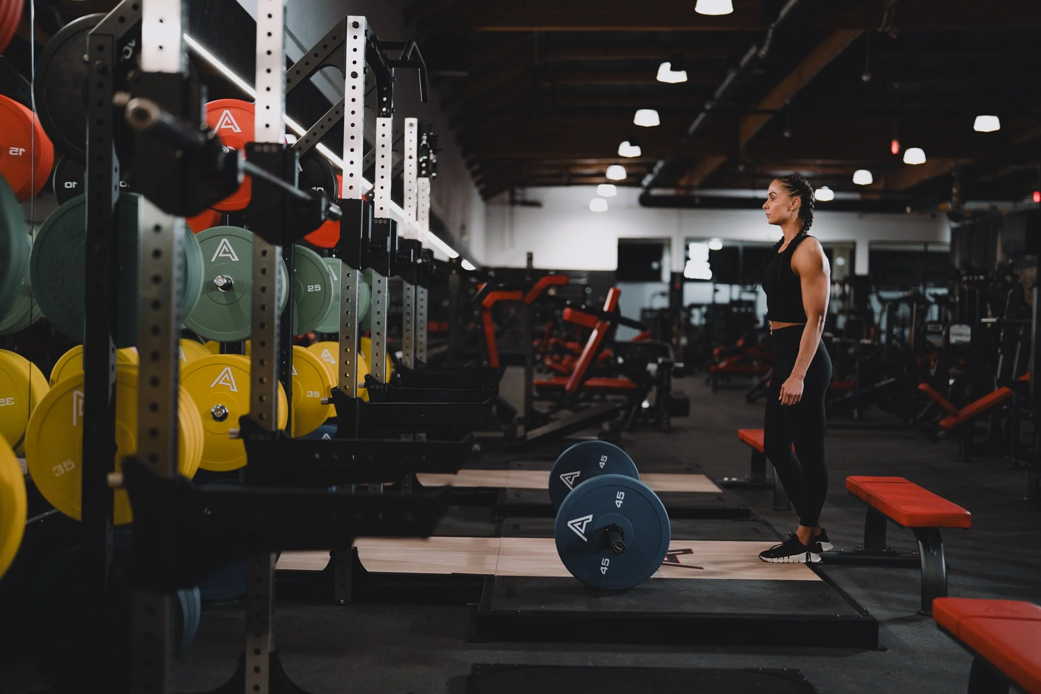 A woman standing in a gym among weightlifting equipment, with colorful weights stored on racks in the background.