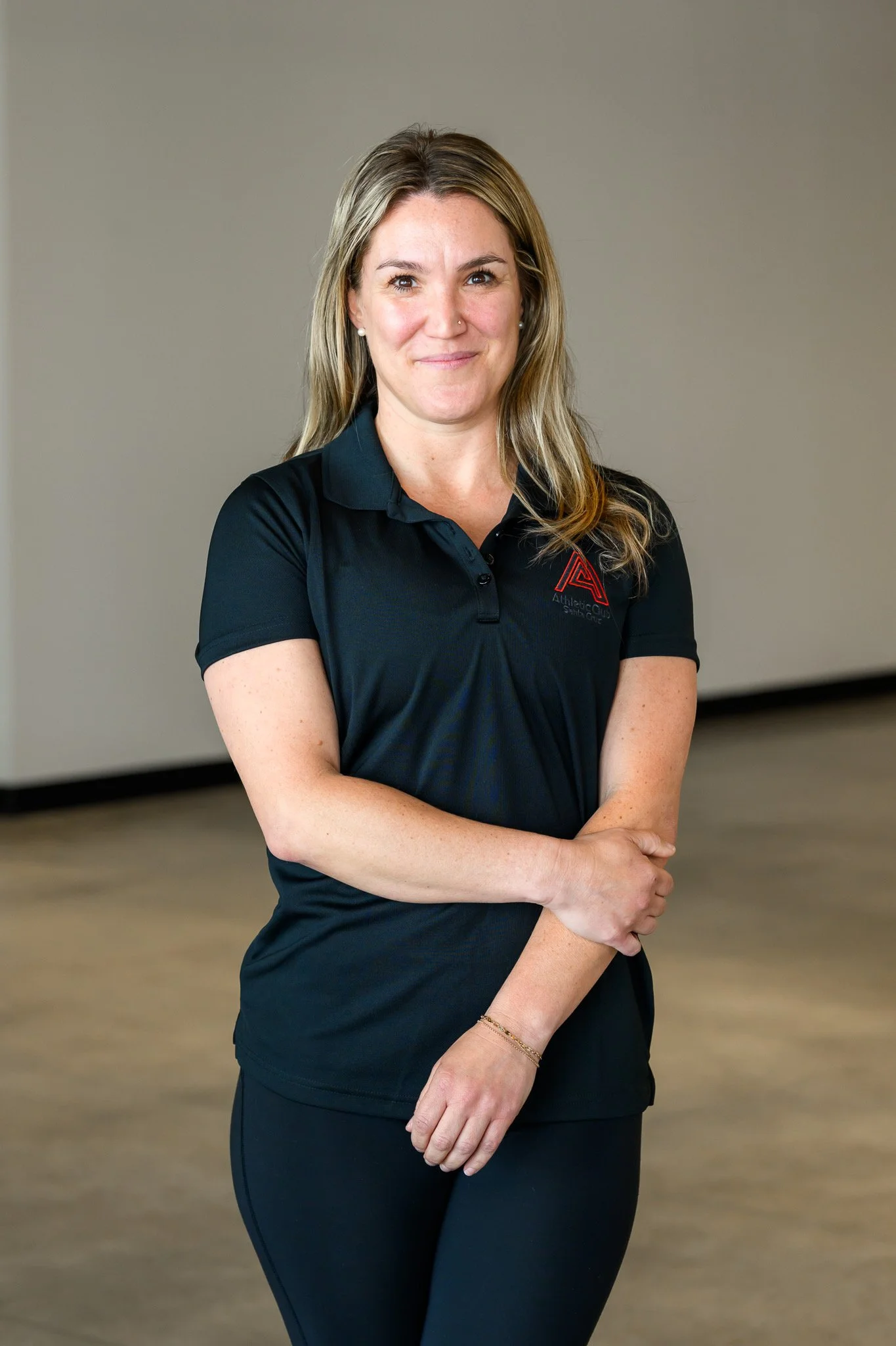 A woman standing indoors wearing a black polo shirt with a red and black logo, smiling with her arms crossed.