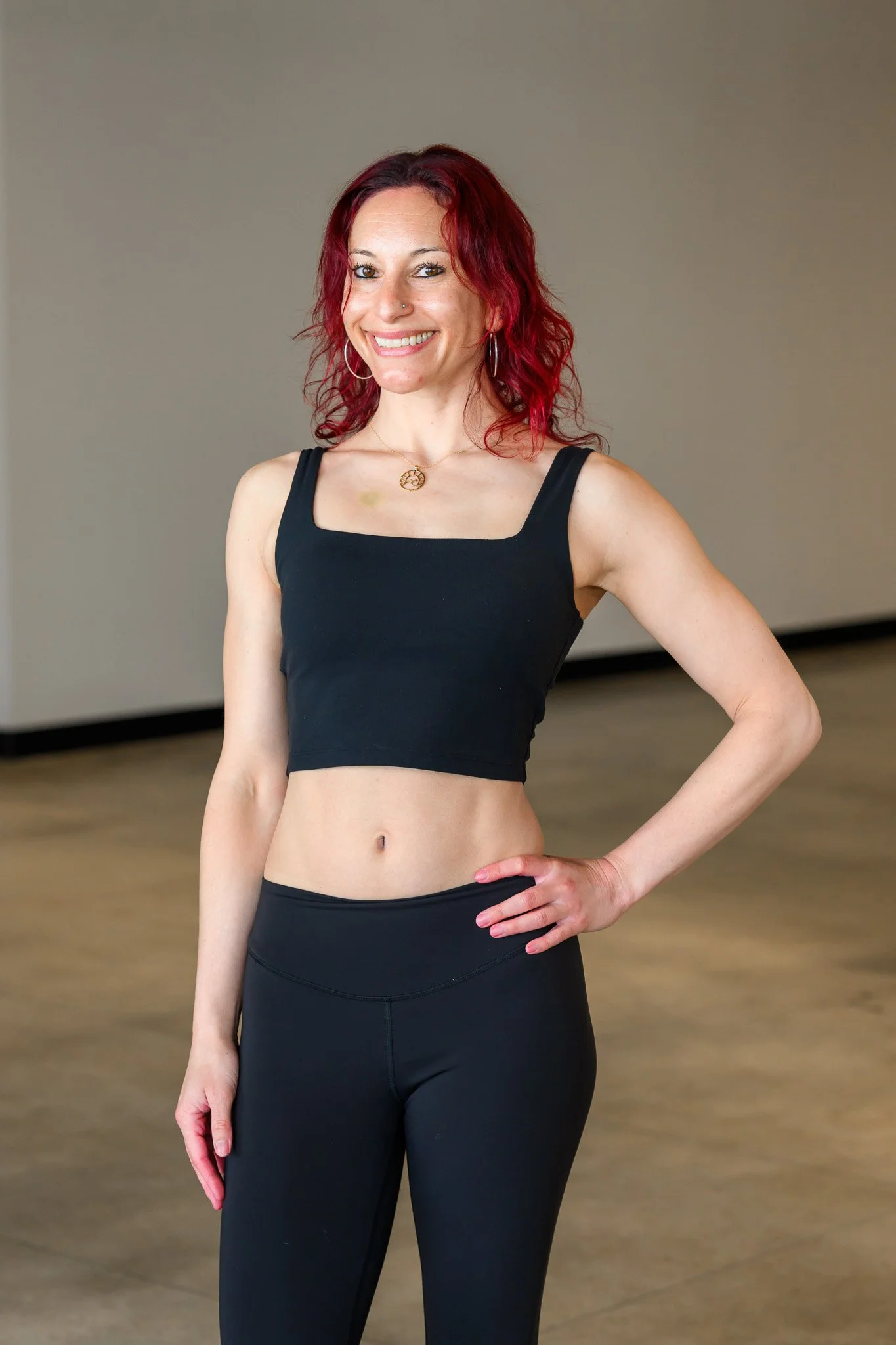 A woman with red, curly hair in workout clothes standing with one hand on her hip, smiling at the camera inside a gym or studio.