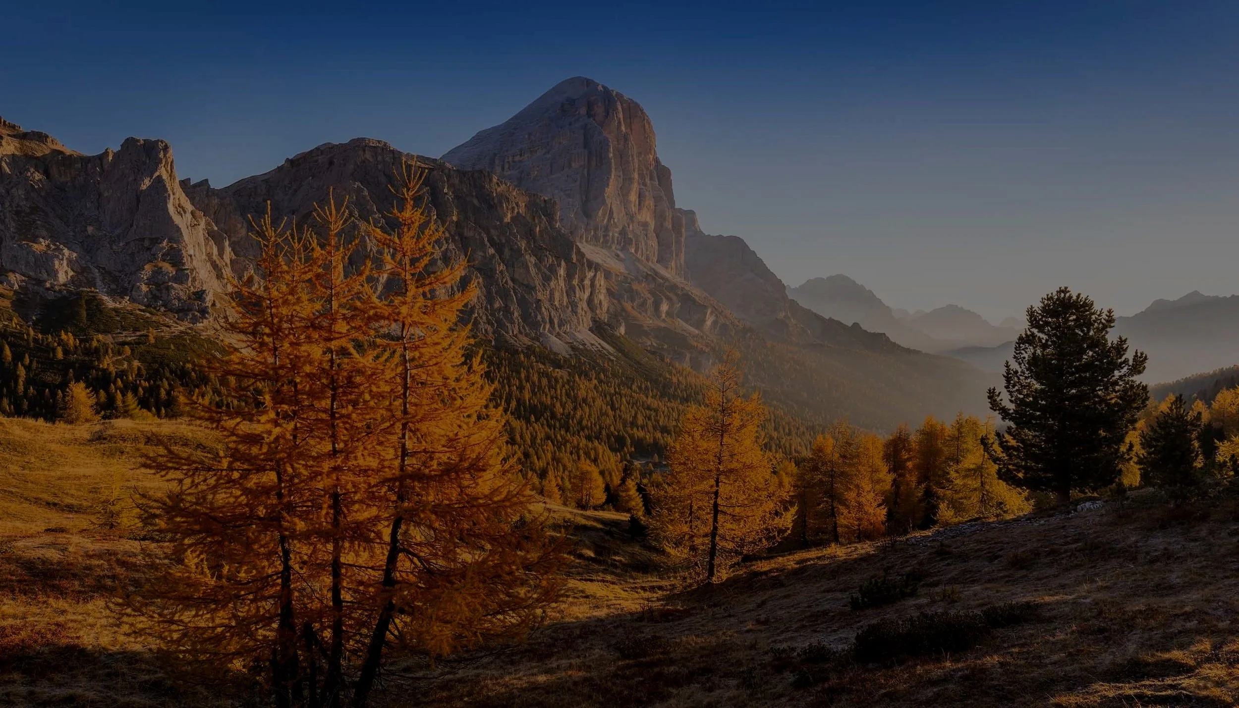 A mountain landscape in fall with orange and yellow trees in the foreground, rugged mountains in the background, and a clear blue sky.