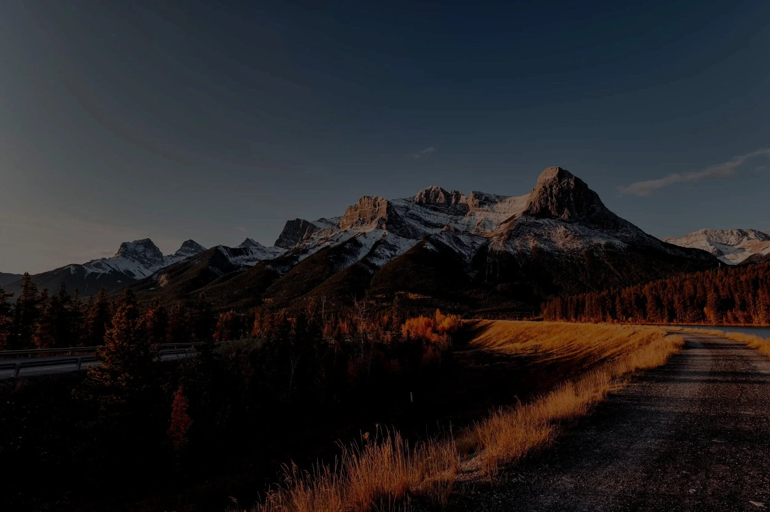 Mountain landscape with snow-capped peaks, a forested base, and a gravel road in the foreground during sunset.