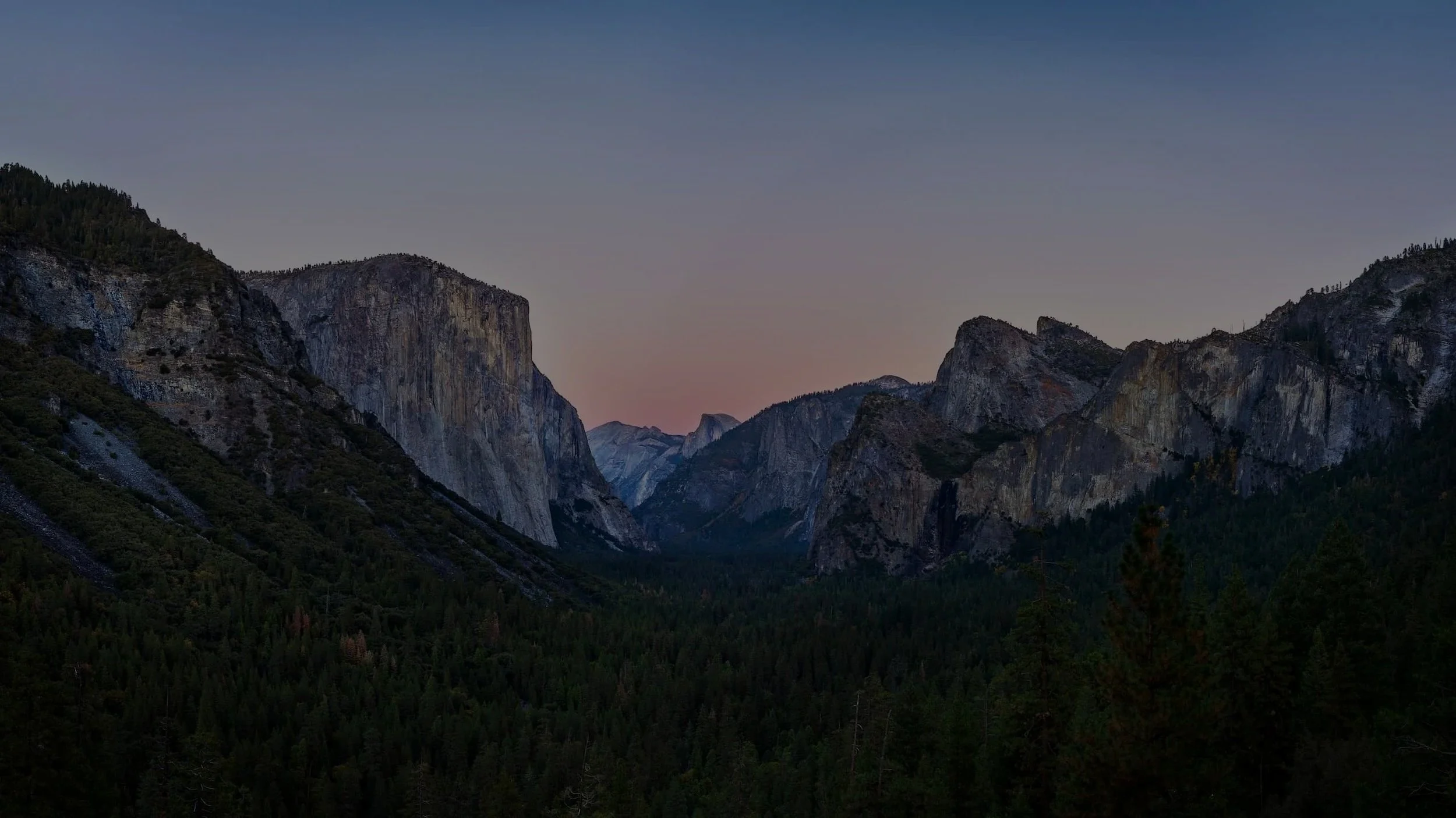 A view of a mountain valley during twilight, with tall cliffs on either side and a dense forest at the bottom.