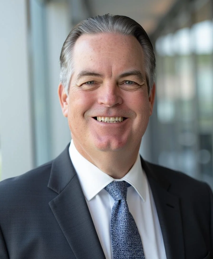 A smiling man in a suit and tie standing in front of a window.
