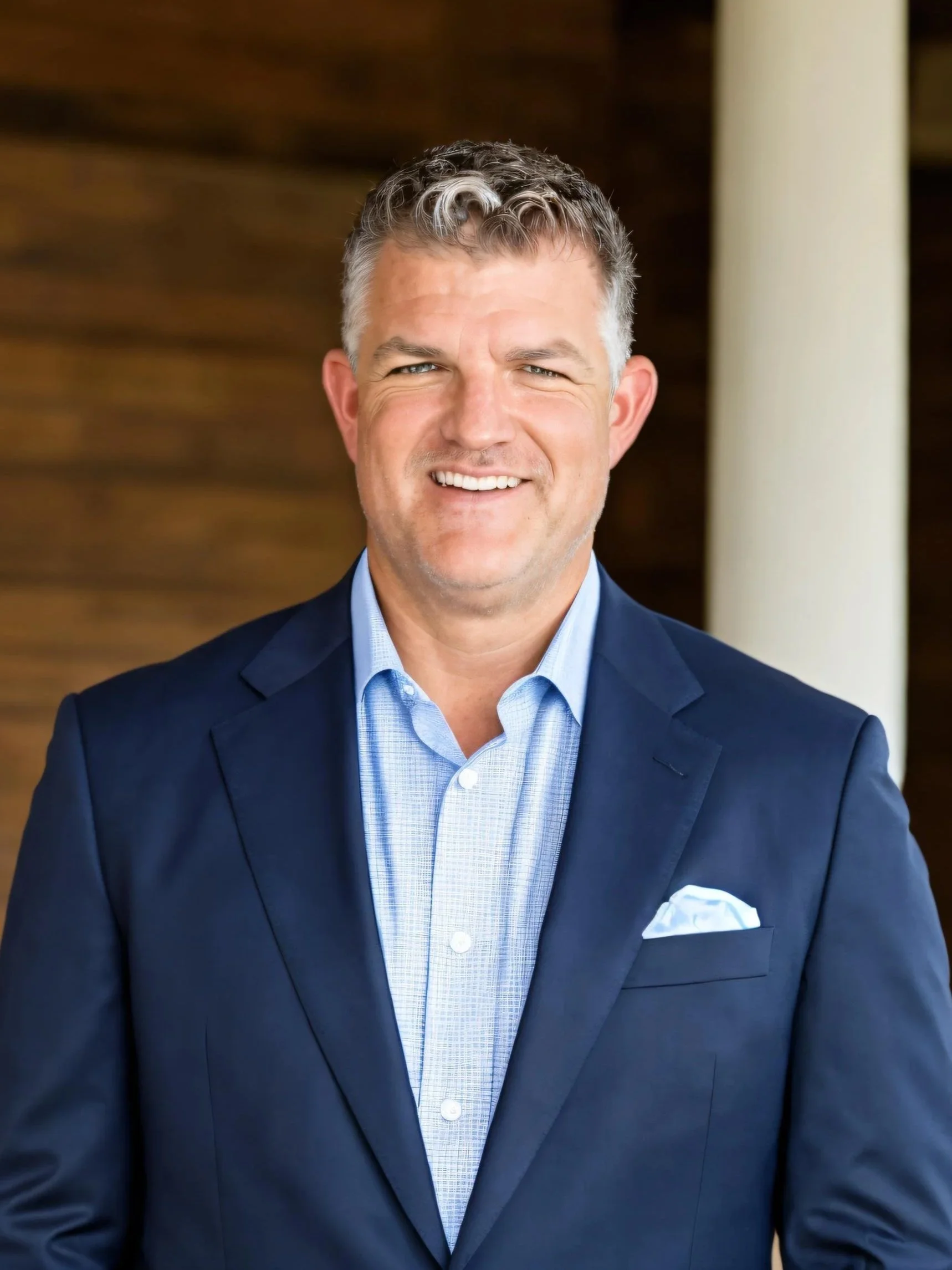 A smiling man in a navy blue suit and light blue dress shirt, standing indoors with a wooden background and a white column.