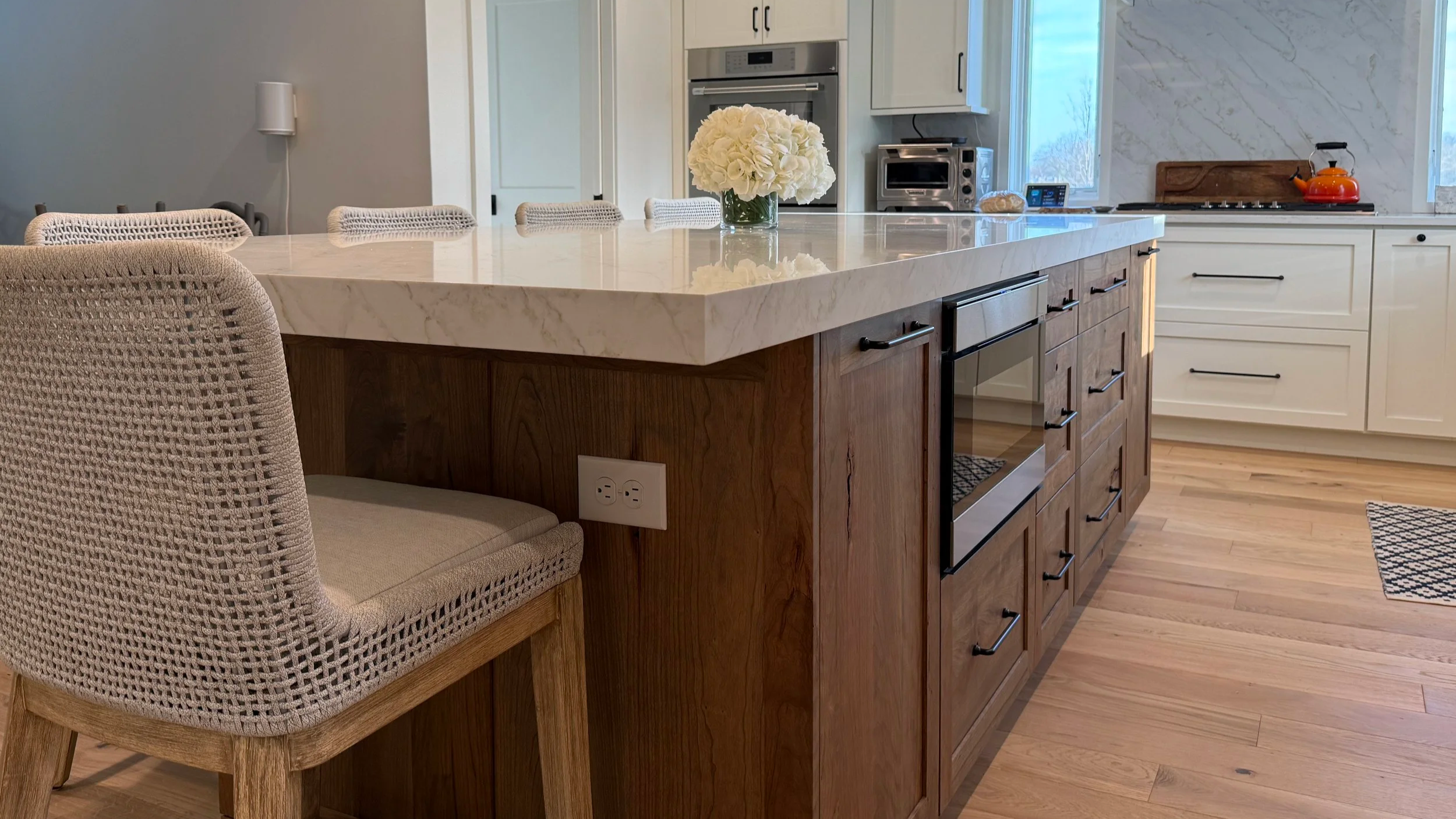 Modern kitchen with a large white marble island, beige upholstered chairs, and wooden cabinetry. A vase with white flowers sits on the island. The kitchen has built-in appliances and a window allowing natural light.